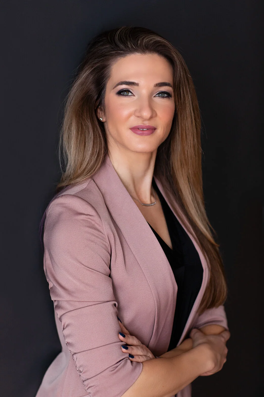 A woman with long brown hair and blue eyes posing against a black background. She is wearing a pink blazer over a black top, with her arms crossed.