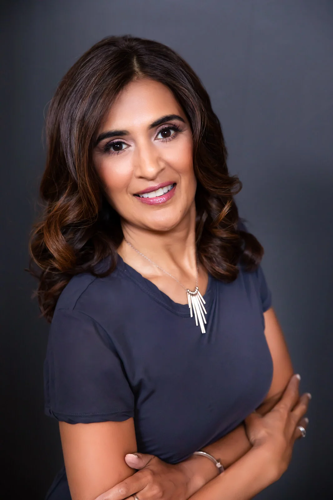 Portrait of a woman with shoulder-length brown hair wearing a navy blue top, smiling, and crossed arms against a dark gray background.