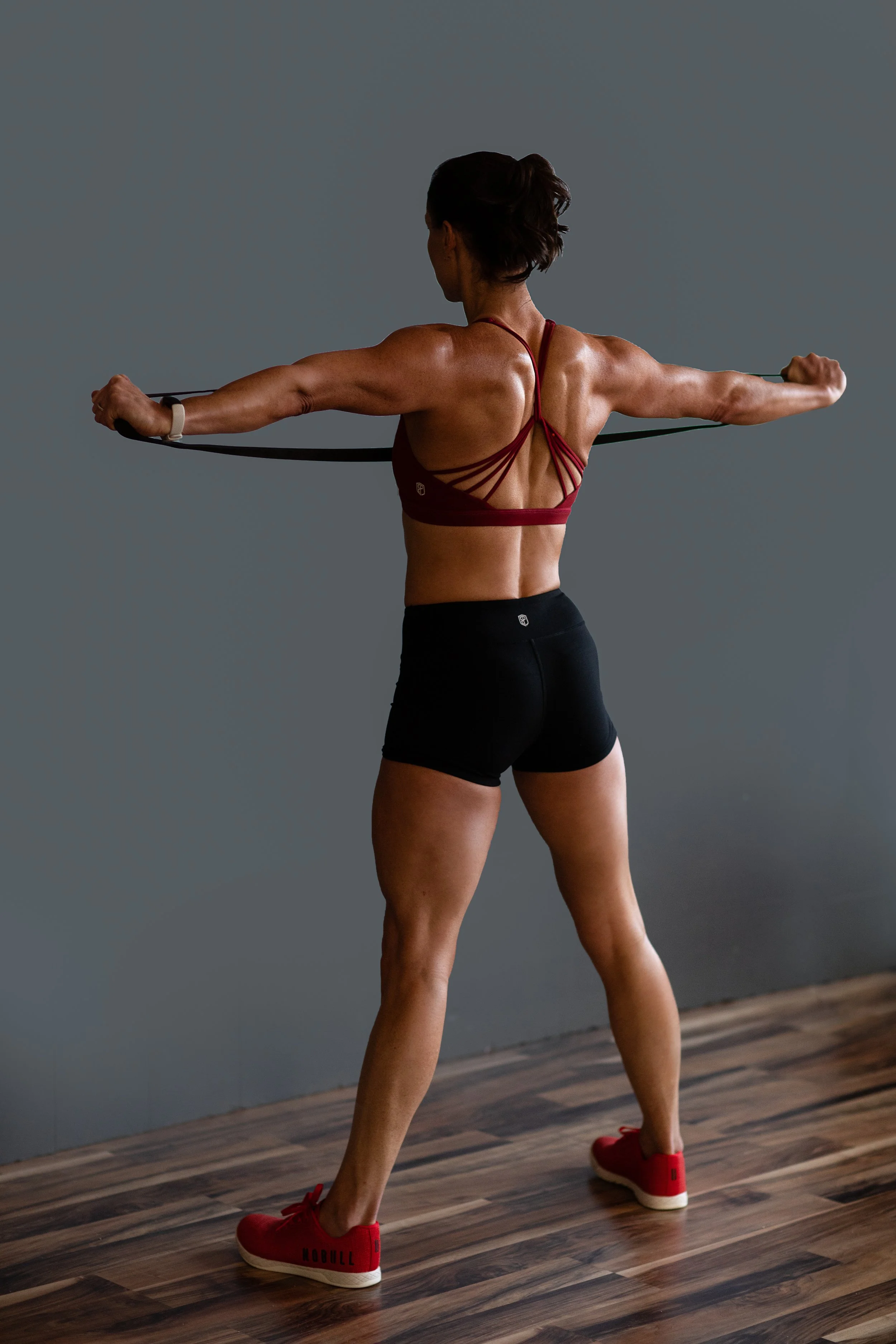 A woman with a muscular back and arms wearing a red sports bra, black shorts, and red sneakers, holding a resistance band extended to her sides in a fitness studio with a wooden floor.