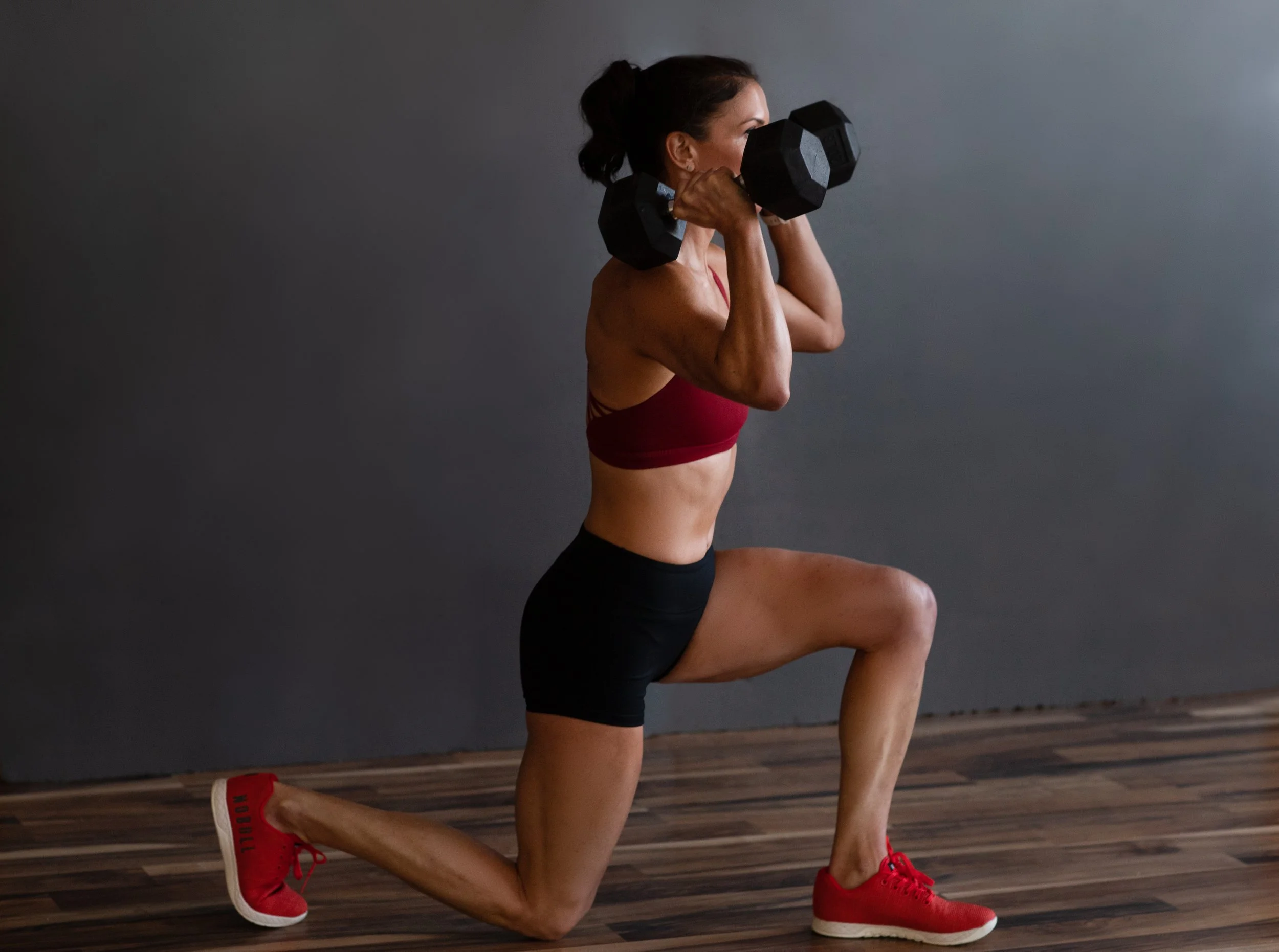 A woman in a workout outfit performs a lunge with a dumbbell held at her shoulders on a wooden floor with a dark gray background.