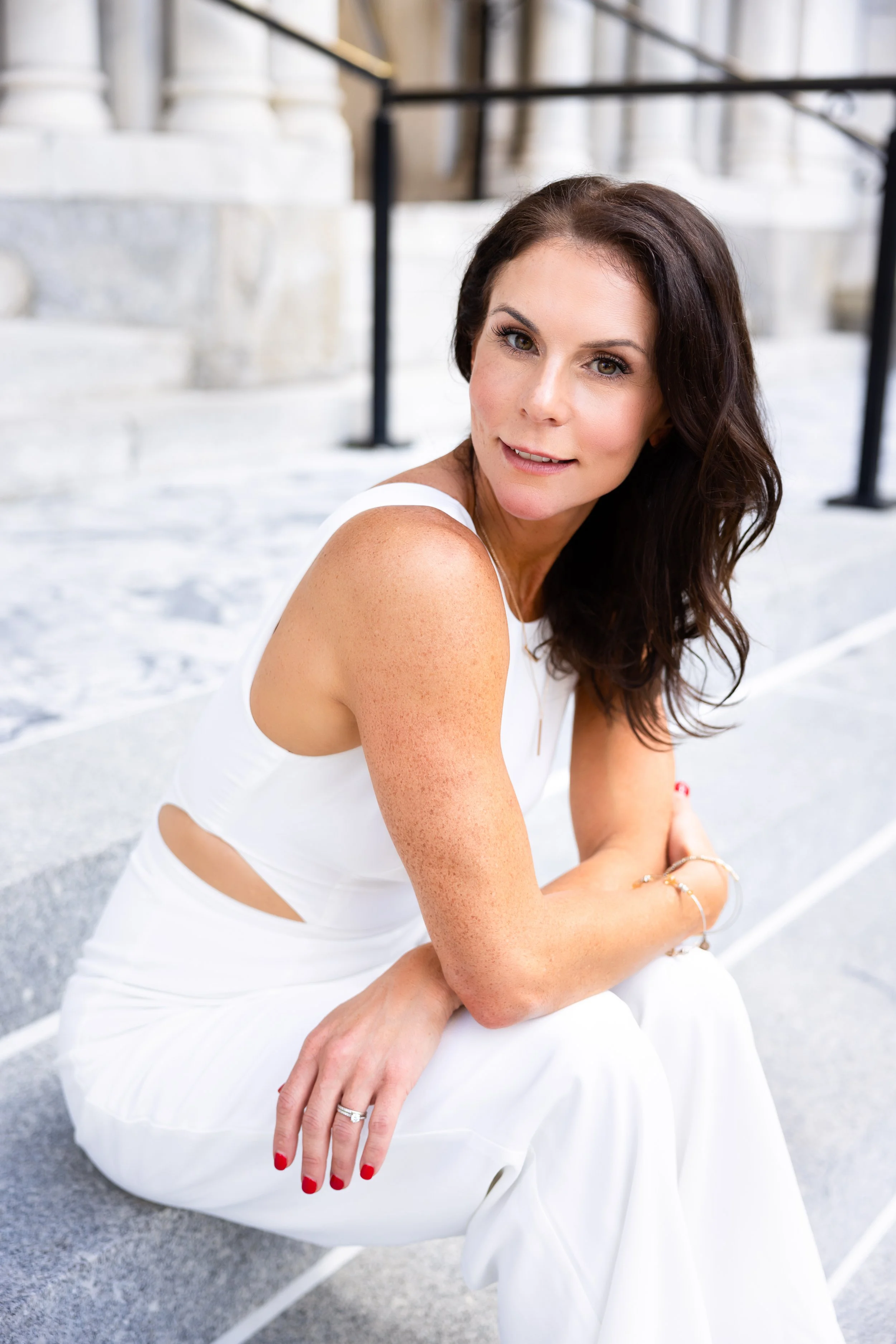 A woman with dark hair wearing a white sleeveless dress and red nail polish, sitting on steps outside of a building, looking at the camera.