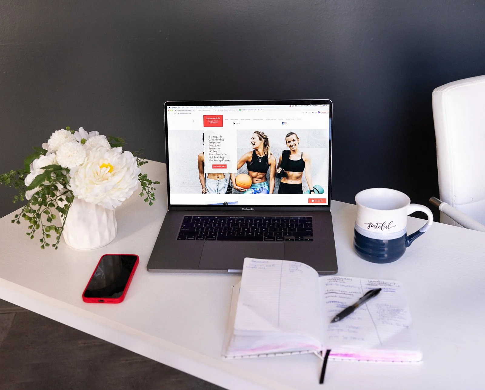 A workspace with a MacBook Pro laptop on a white desk displaying a fitness website with two women in workout clothes. To the left of the laptop, there is a white vase with white flowers and green foliage, and a red-edged smartphone. To the right, the