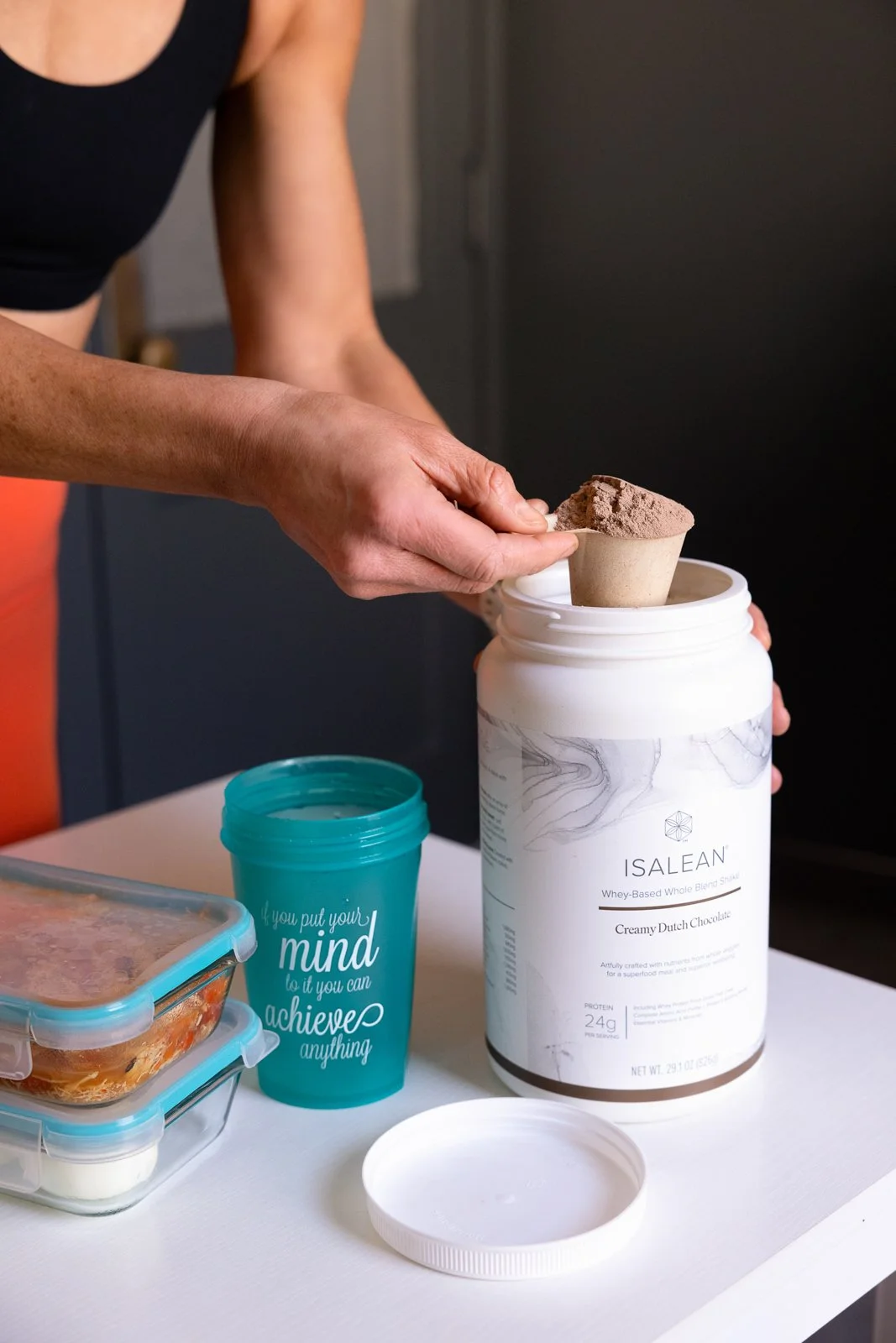 A person scooping protein powder into a container of whey protein powder on a white surface, with food containers and a motivational quote cup nearby.
