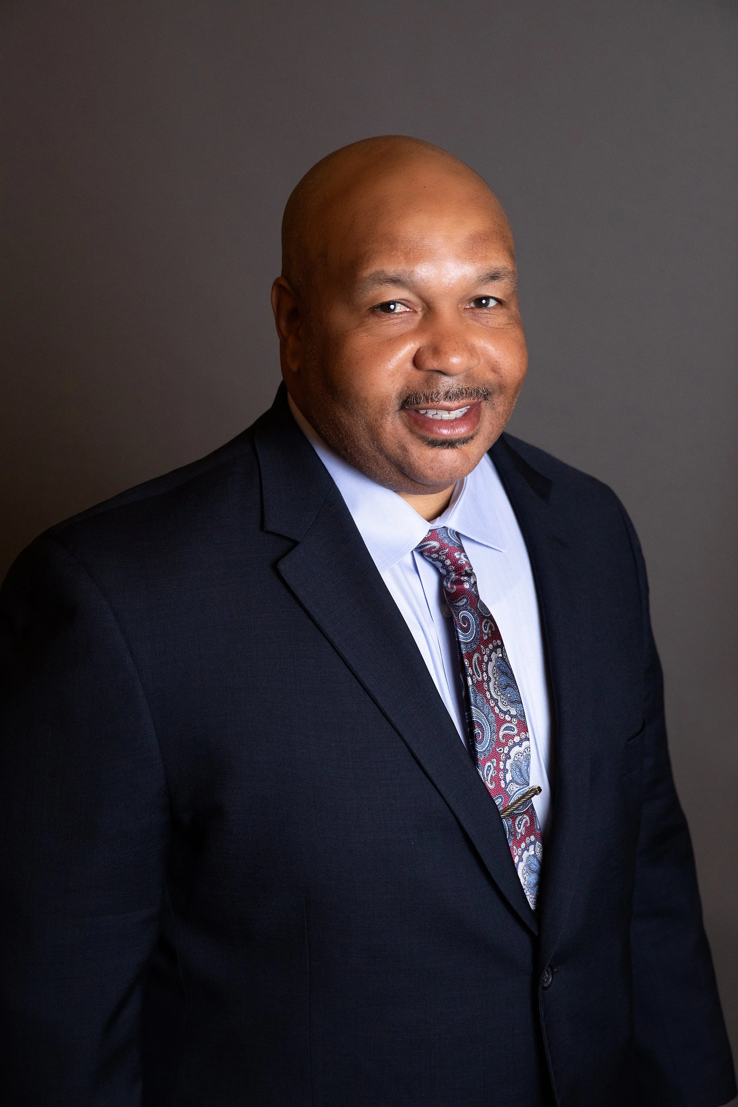 A man in a dark suit, light blue shirt, and a paisley tie, smiling at the camera against a plain dark background.
