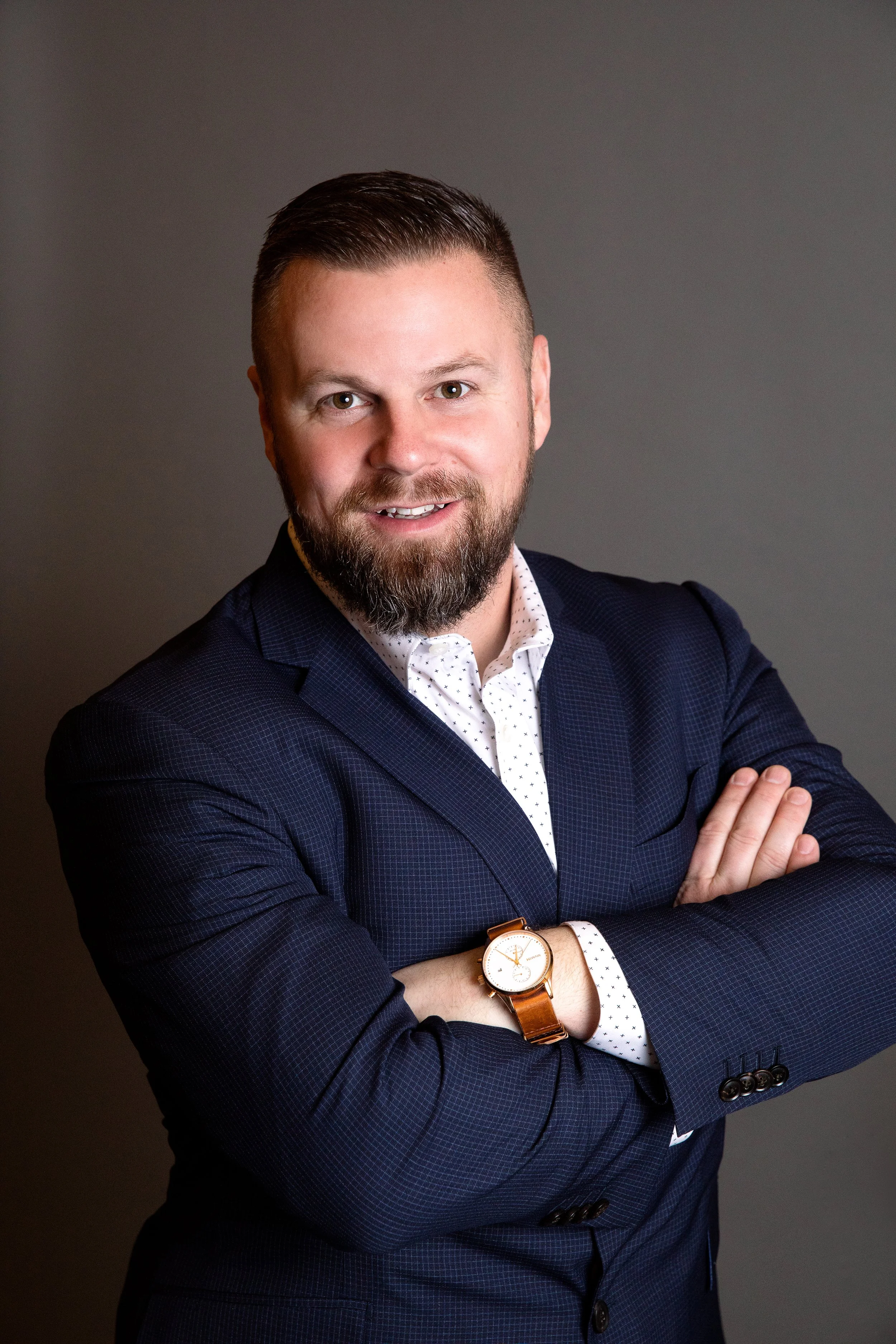 A man with a beard and short hair smiling, wearing a dark blue suit, white dotted shirt, and a watch, standing with arms crossed against a gray background.