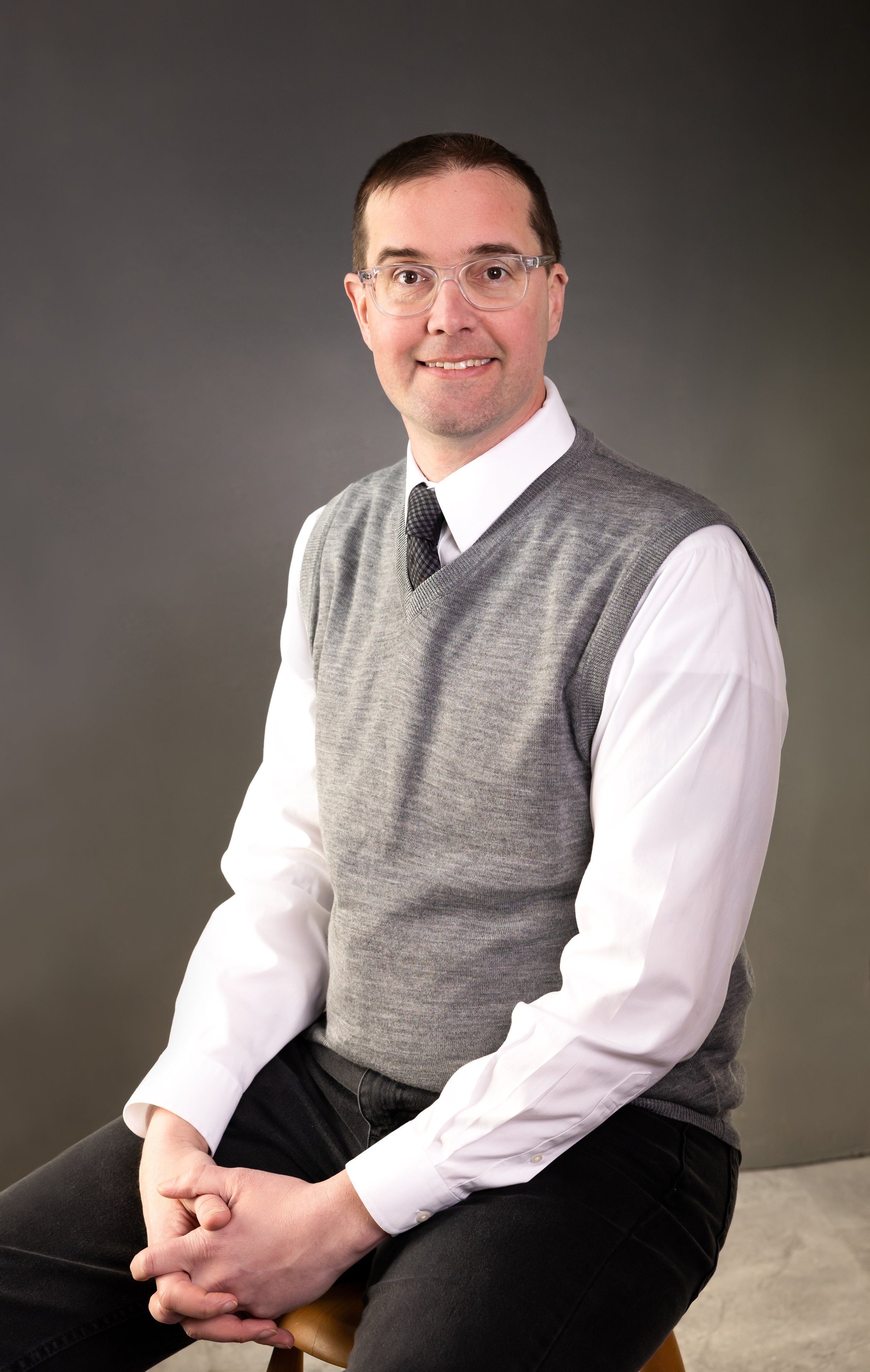 A man wearing glasses, a white shirt, a gray sweater vest, and a dark tie, sitting on a stool against a gray background, smiling at the camera.