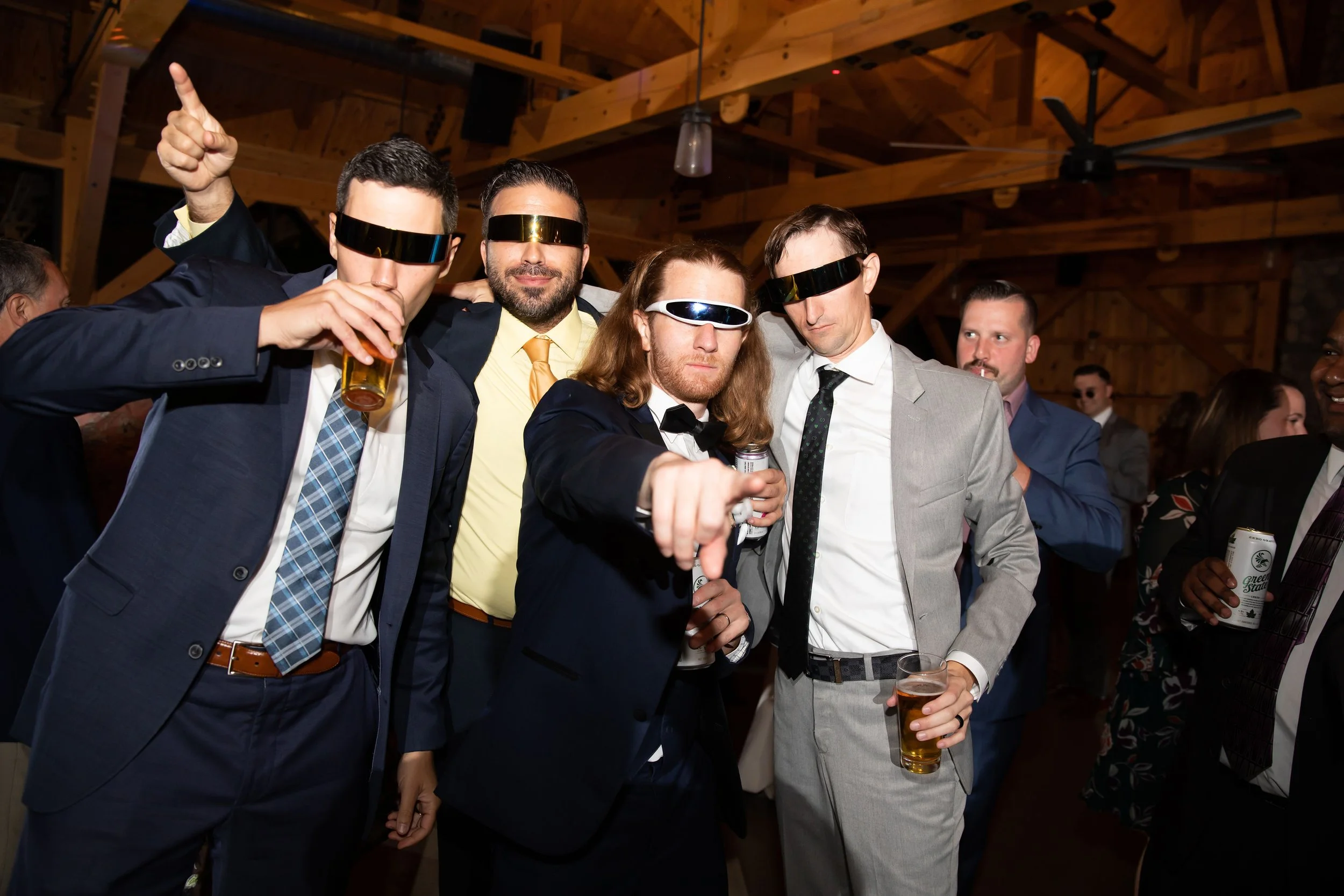 Group of men in suits wearing night-vision goggles at a party, holding drinks, in a wooden venue.
