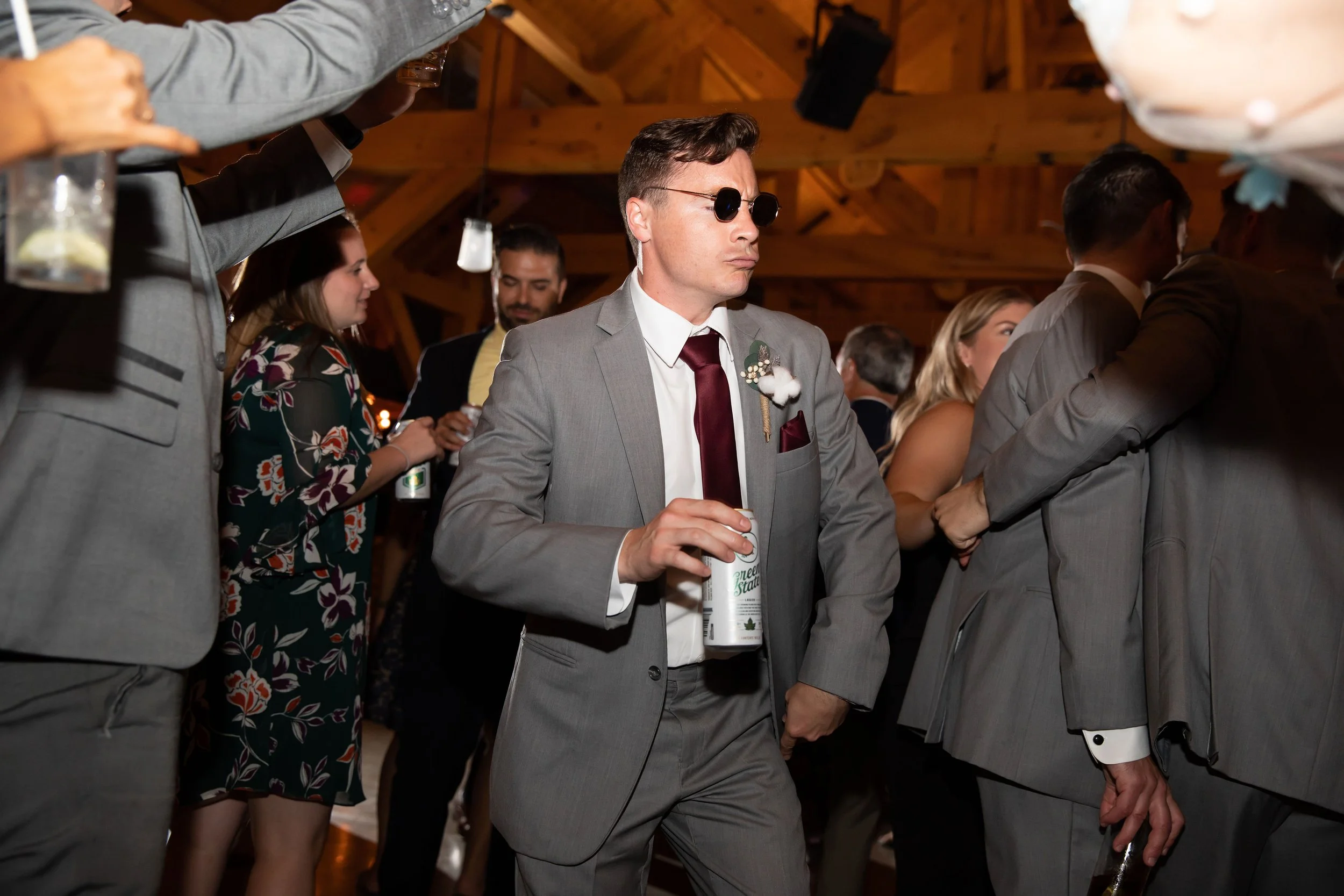 Man in gray suit and sunglasses holding a drink at a wedding reception in a wooden venue