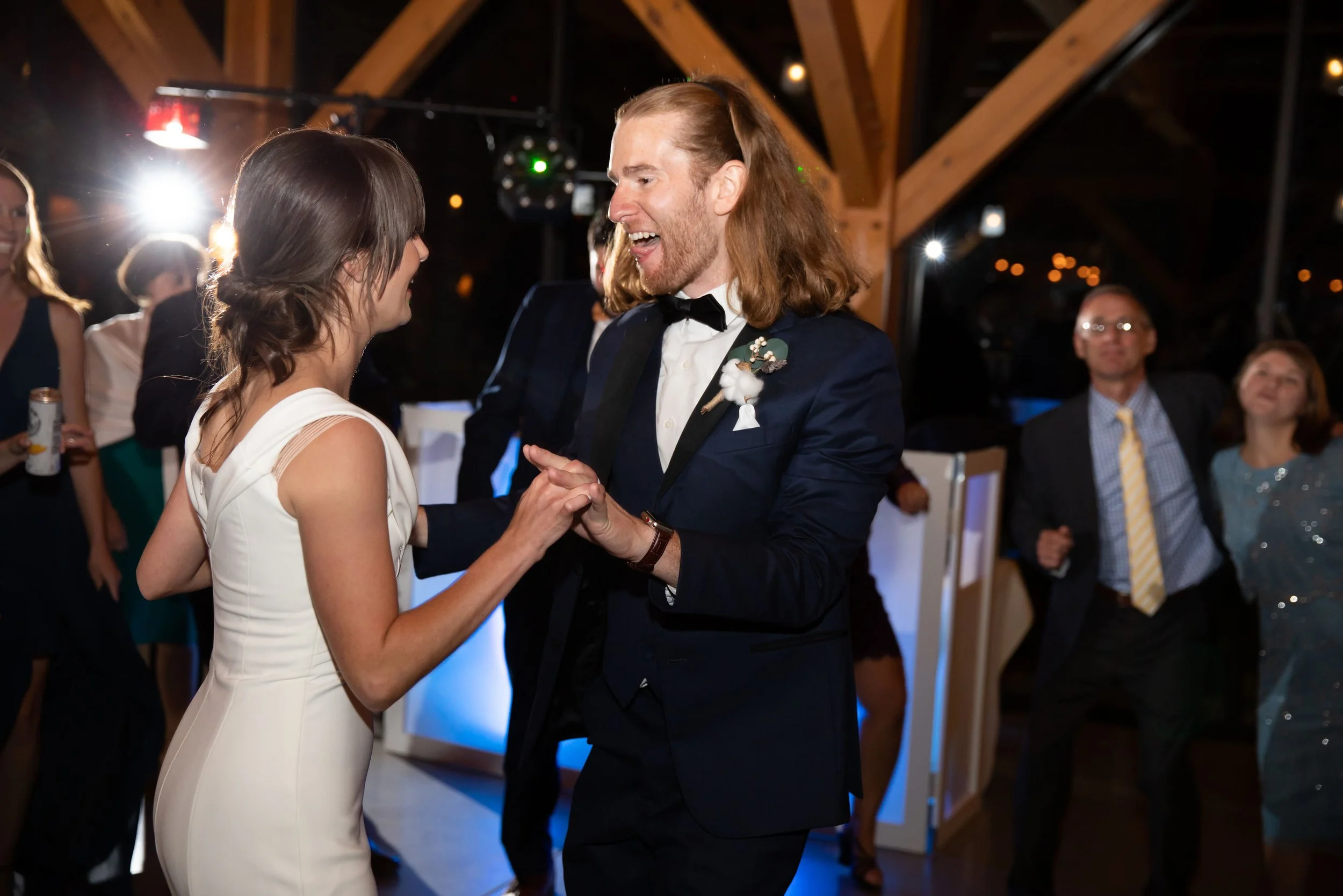 A couple dancing at a wedding reception. The man has long hair and is wearing a dark tuxedo with a boutonnière, smiling at the woman in a white dress. People are dancing and celebrating in the background.