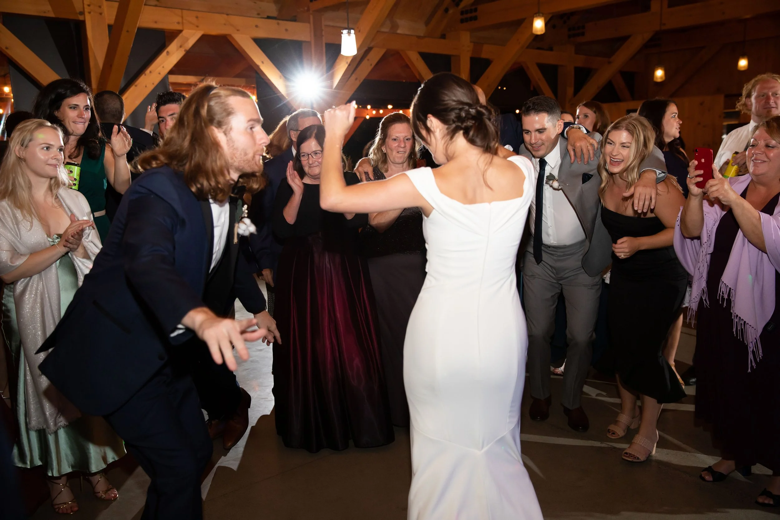 A bride in a white wedding dress and a groom in a gray suit dance together at a wedding reception, surrounded by smiling guests in a wooden hall.