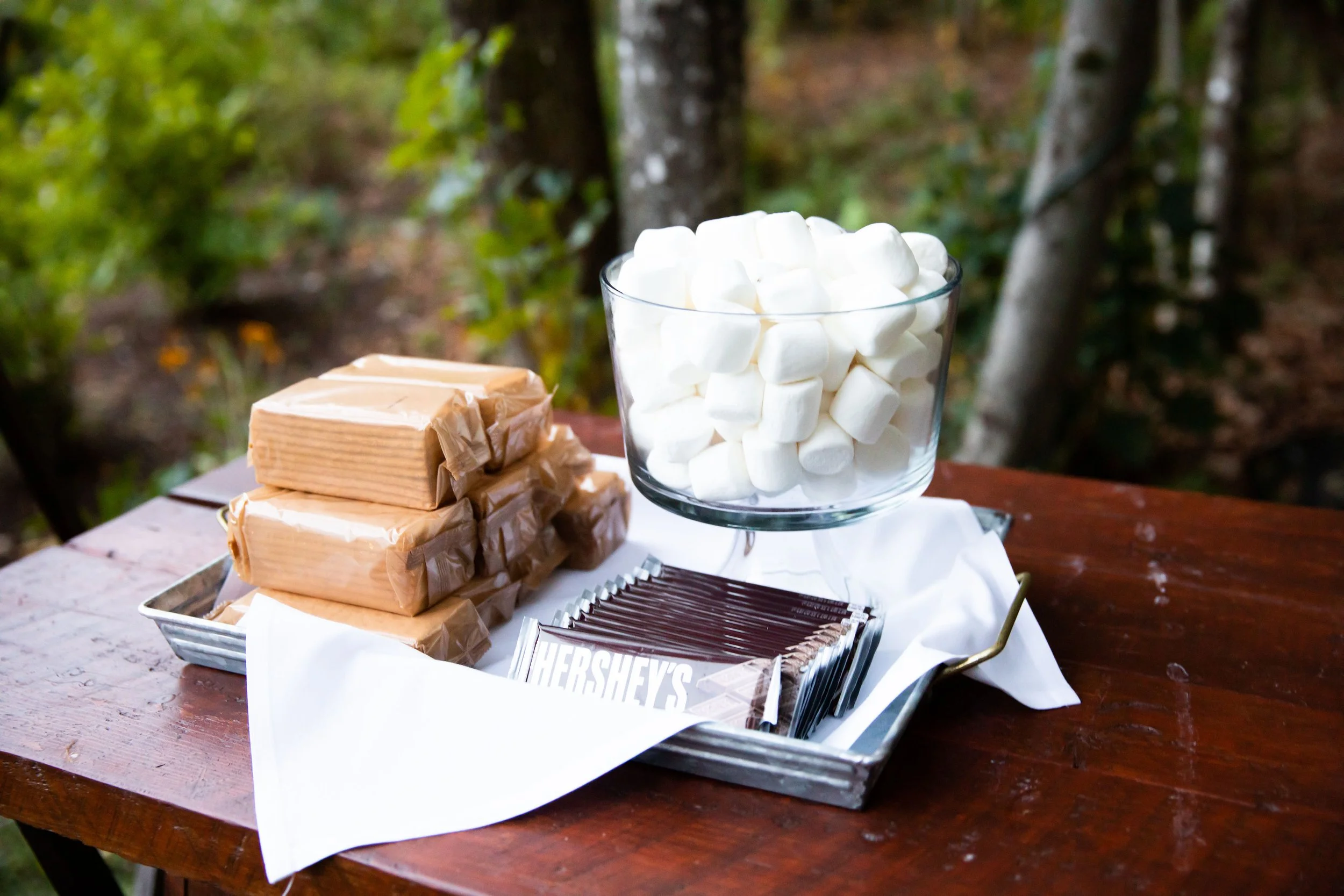 A tray with stacked caramel bars, a bowl filled with mini marshmallows, and packets of Hershey's chocolate on a wooden table outdoors.