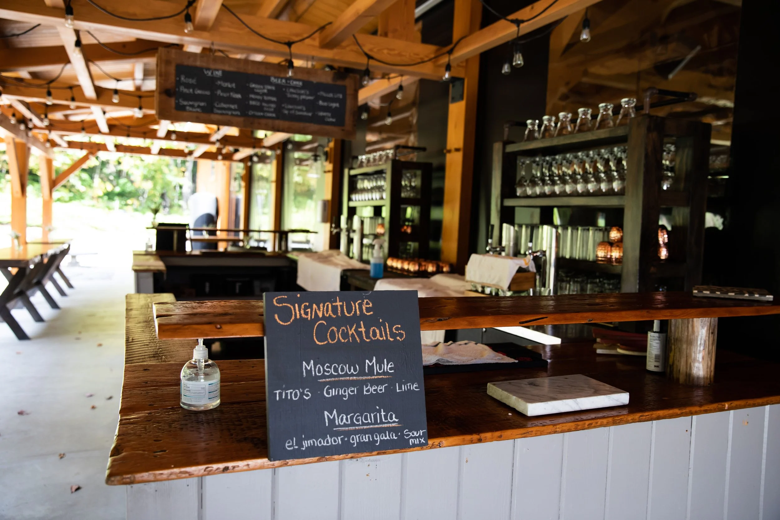A bar counter with a handwritten chalkboard sign listing signature cocktails, including Moscow Mule, Tito's Ginger Beer Lime, and Margarita, in a rustic, open-air setting.
