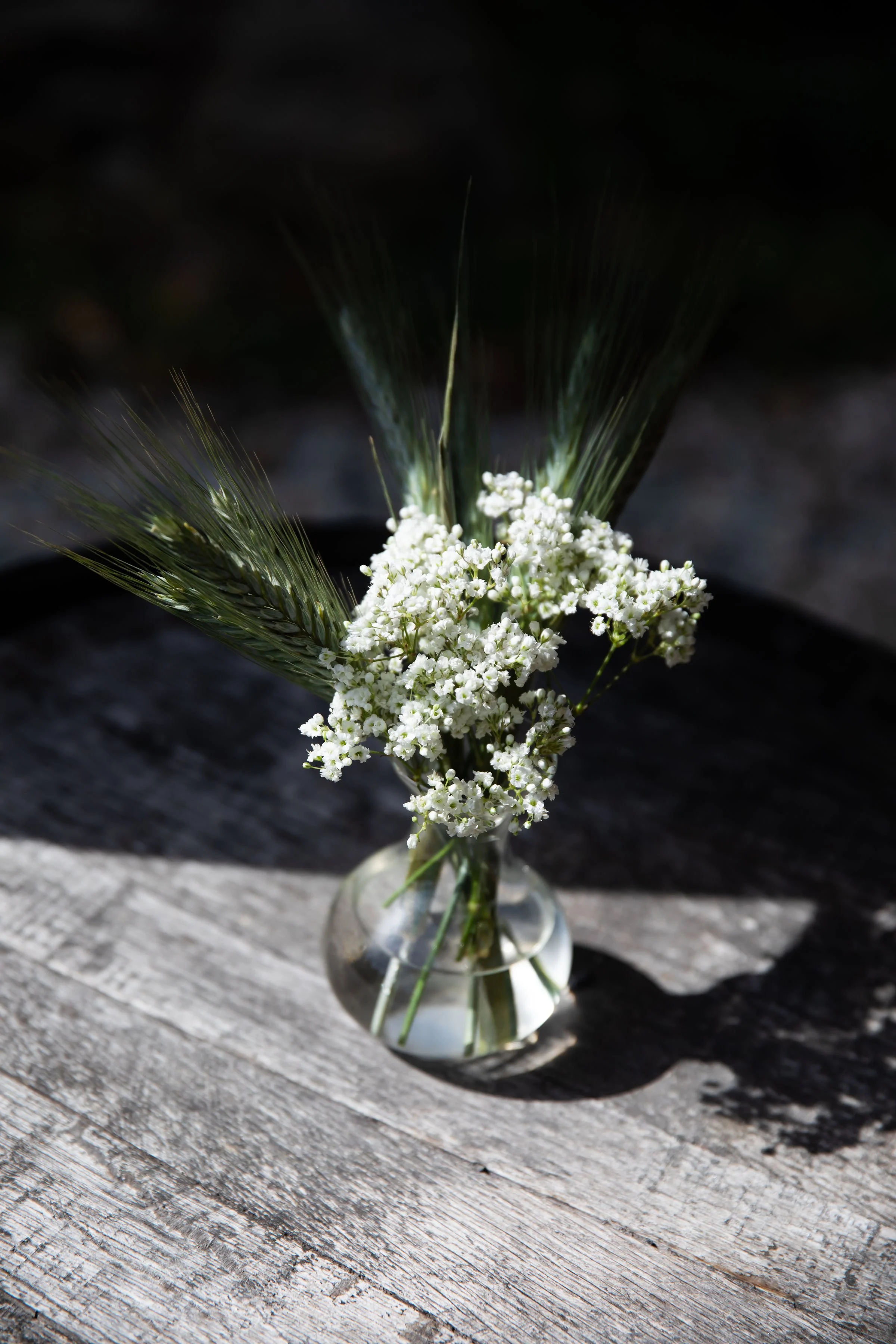 A small glass vase with white flowers and green wheat stems on a weathered wooden surface.