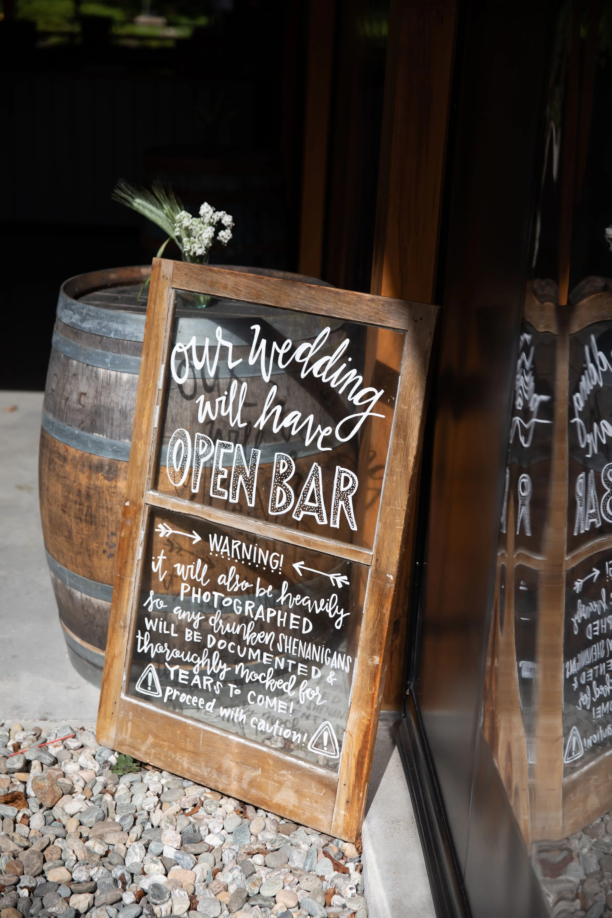 A wooden framed glass signboard with handwritten text announcing an open bar at a wedding. The sign also warns that it will be heavily photographed and that drunken shenanigans will be documented, advising caution.