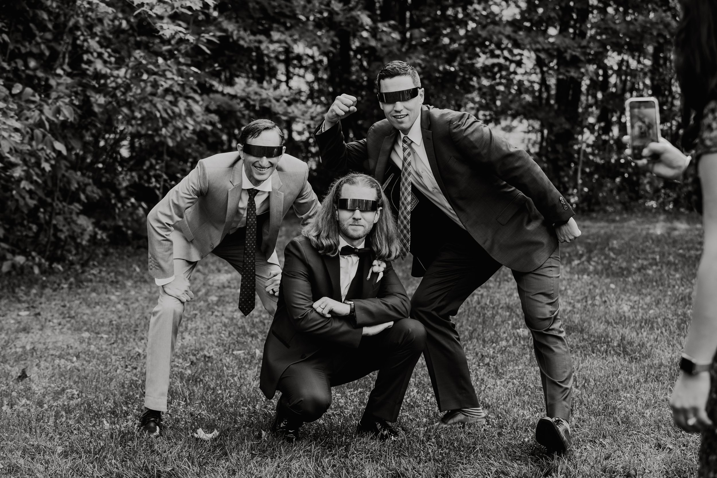 Four men in suits wearing virtual reality glasses posing playfully outdoors on grass with leafy trees in the background, one crouching and the others standing, with a person on the right taking a photo.