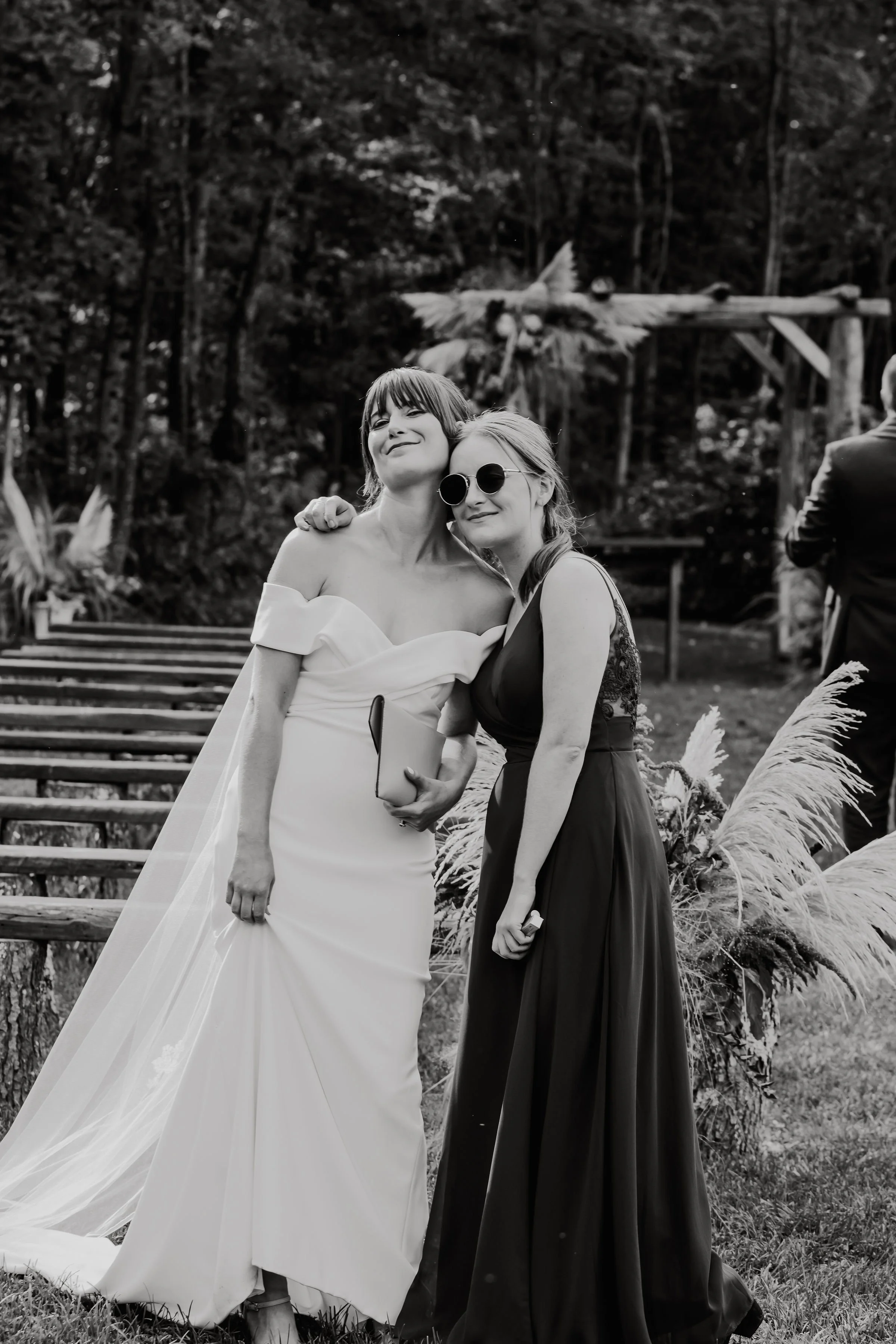 Two women, one in a wedding dress, embrace and smile at an outdoor wedding ceremony, with wooden steps and decorative foliage in the background.
