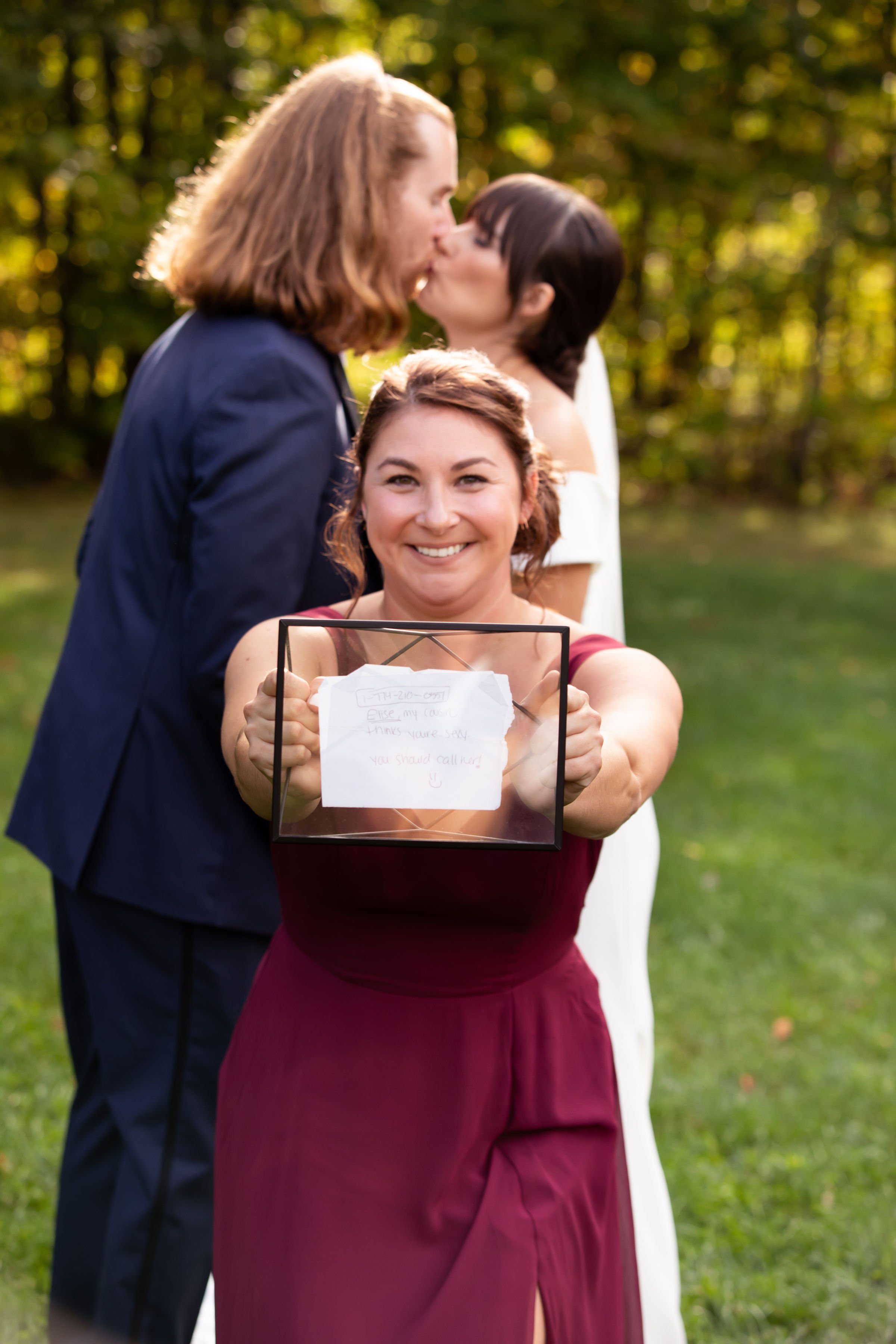 A woman in a burgundy dress holding and presenting a note in a glass frame, smiling, with a wedding ceremony of two women in the background, one kissing the other, outdoors with trees and sunlight.
