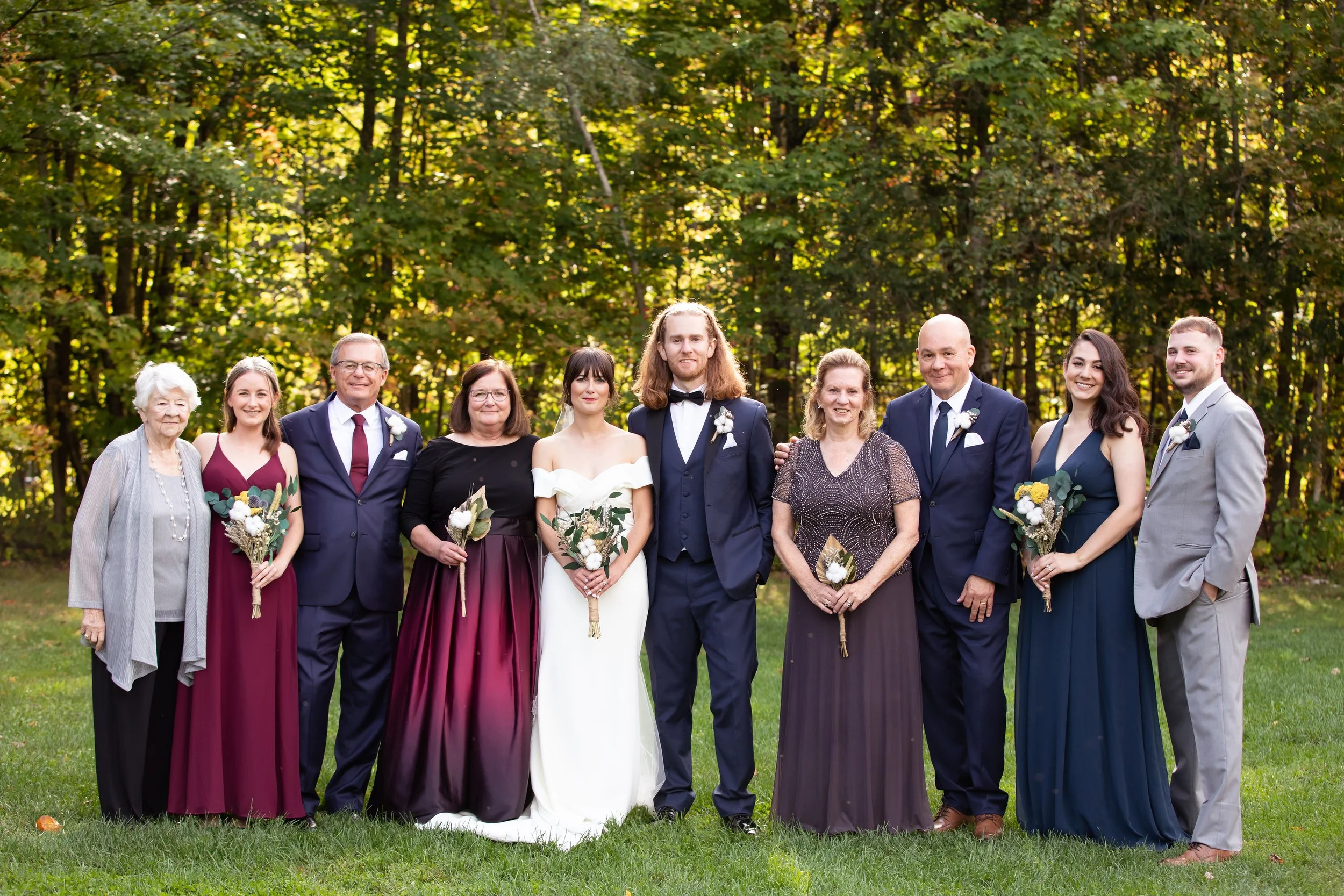 A family group at a wedding outdoor in front of a wooded background, with women in formal dresses and men in suits, some holding bouquets of flowers.