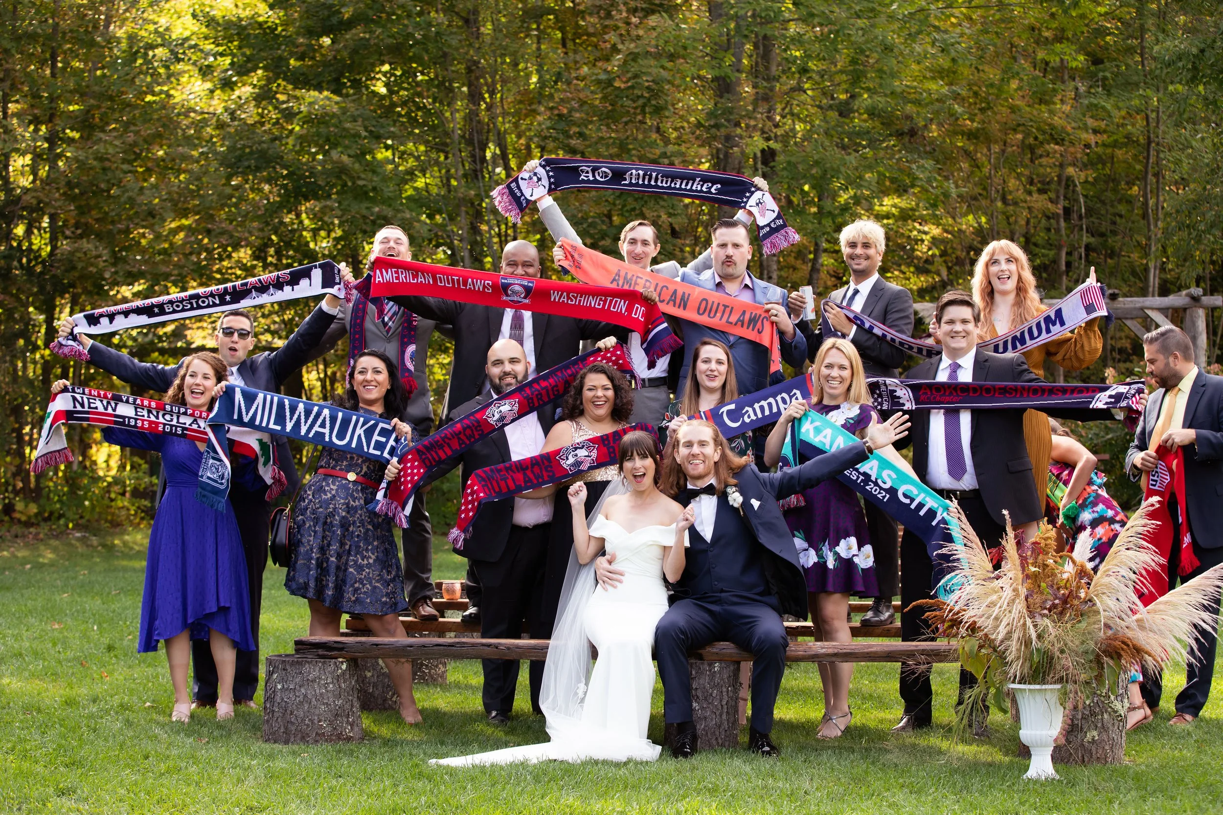 A group of people celebrating outdoors, dressed in formal and casual clothes, holding up colorful scarves with team names and locations, with a bride and groom sitting in front, all smiling and happy.