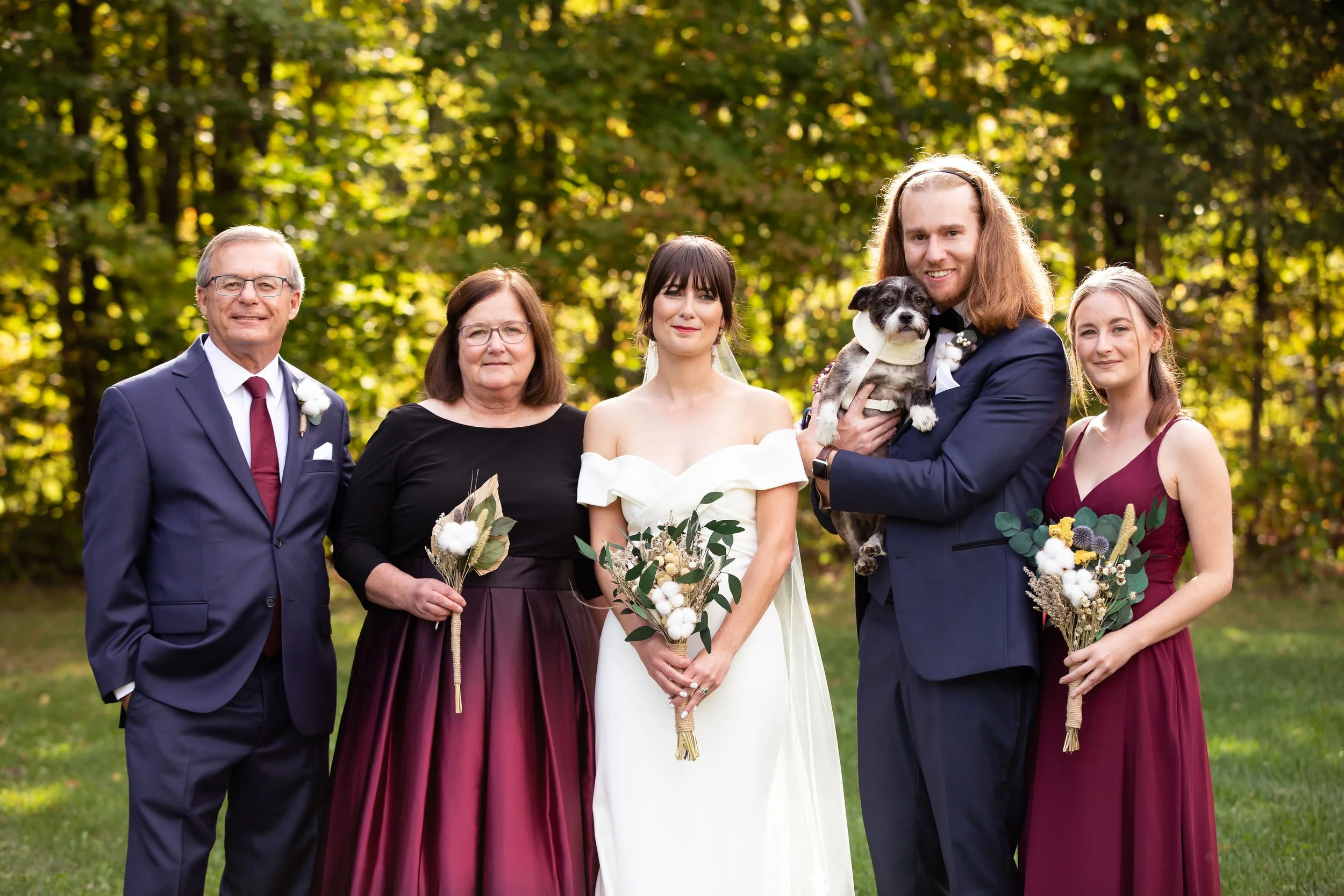 Group of six people, including a bride and groom, standing outdoors with a man, a woman, and two young women, all dressed formally for a wedding, with a dog held by the groom, in a lush green setting.