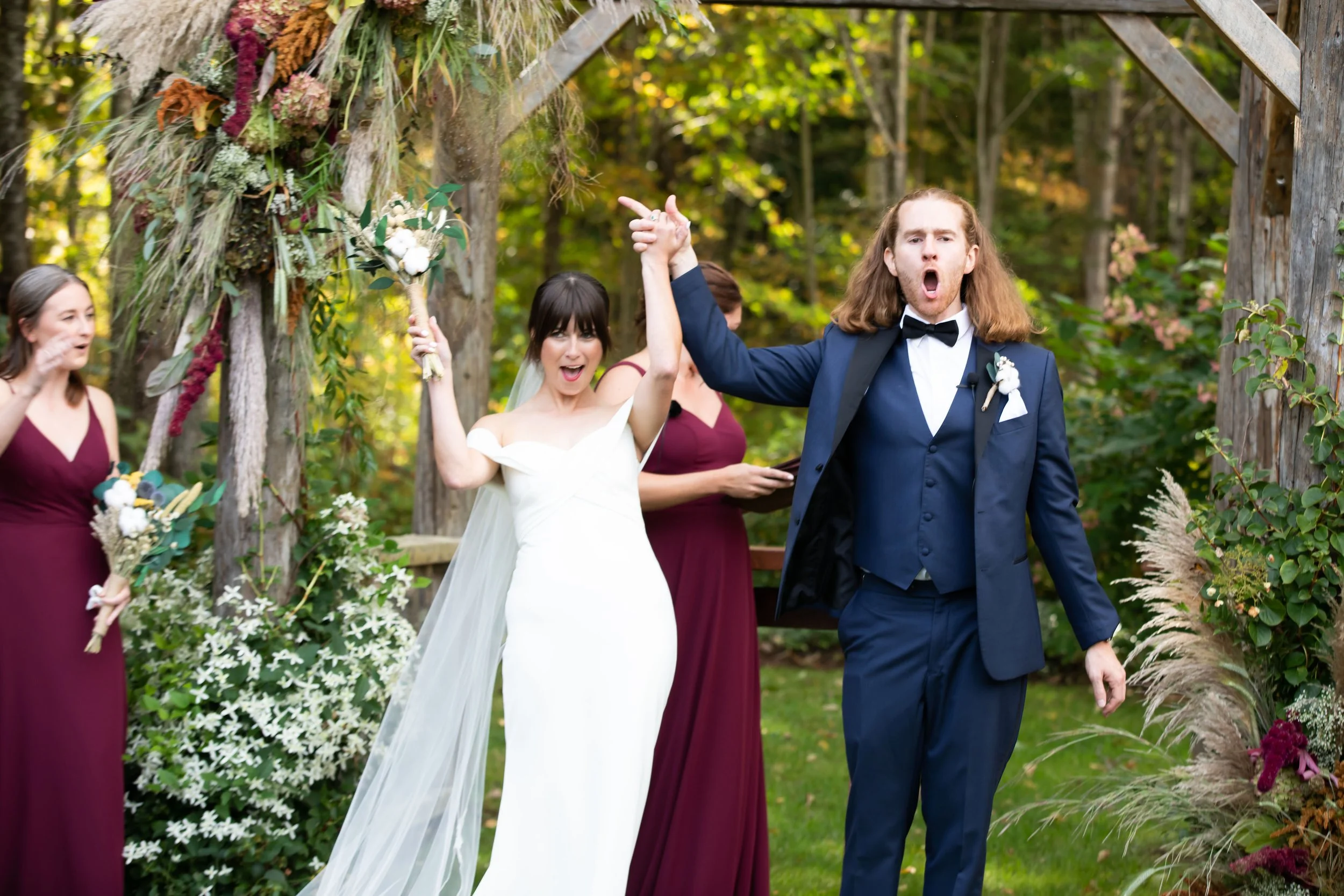 A wedding celebration outdoors with the bride and groom holding hands, surrounded by bridesmaids. The bride is in a white gown and holding a bouquet, the groom is in a navy blue suit and bow tie, all appearing joyful.
