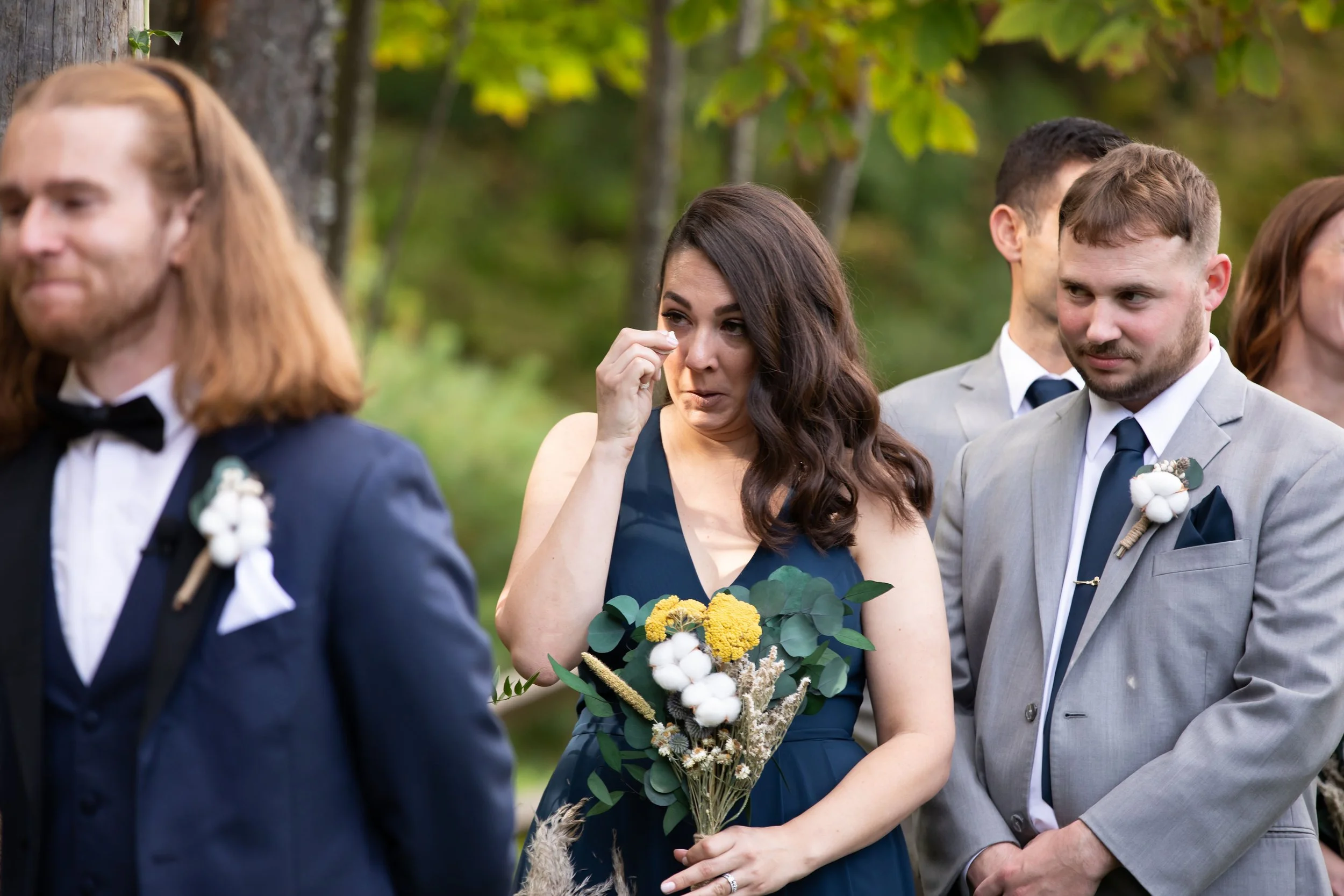 A woman in a blue dress holding a bouquet of flowers wipes away a tear during an outdoor wedding ceremony, with several men in formal suits and ties nearby.