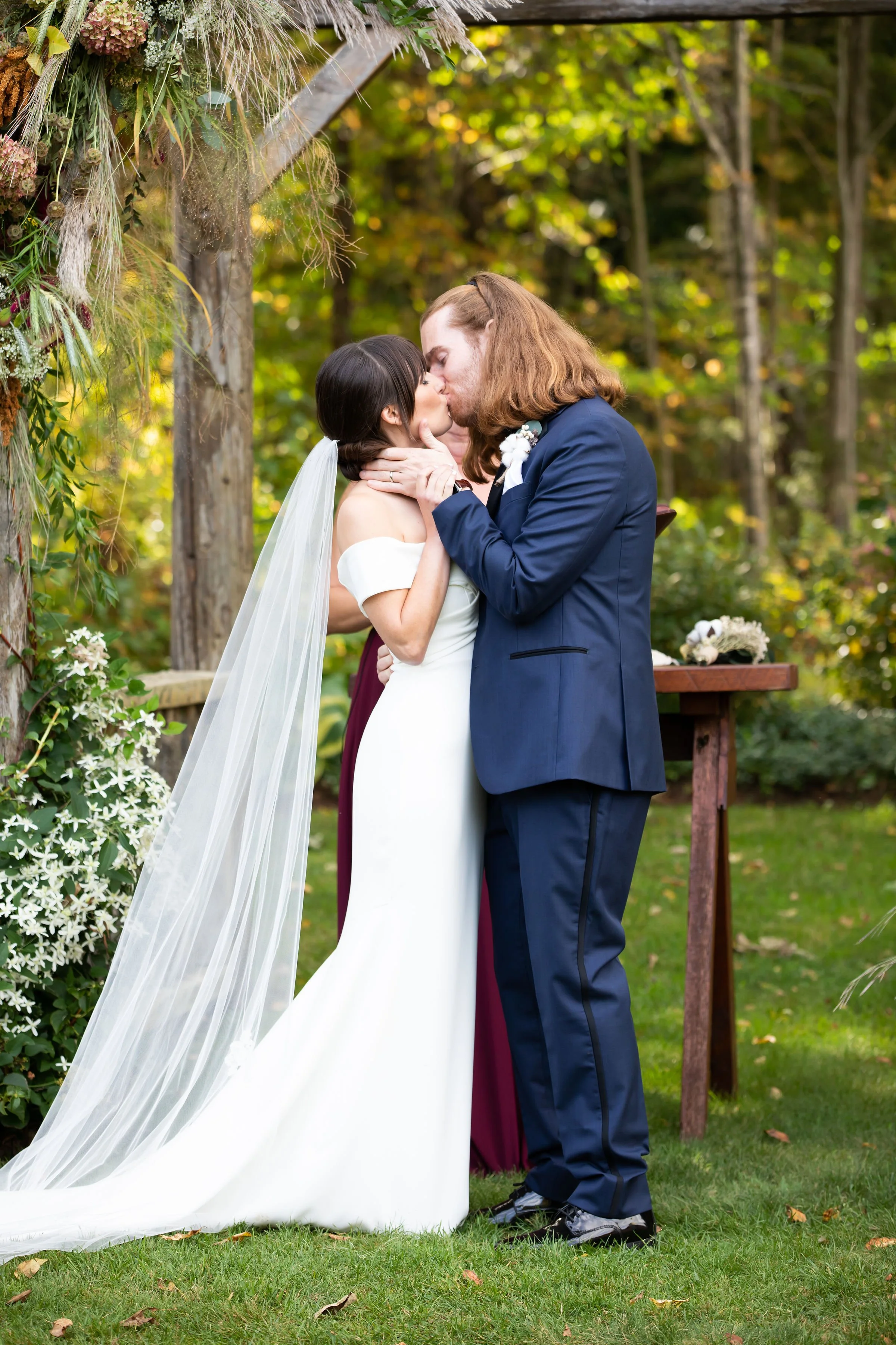A couple sharing their wedding kiss during their outdoor wedding ceremony, with the bride in a white gown and veil and the groom in a navy suit, under a decorated arch surrounded by greenery.
