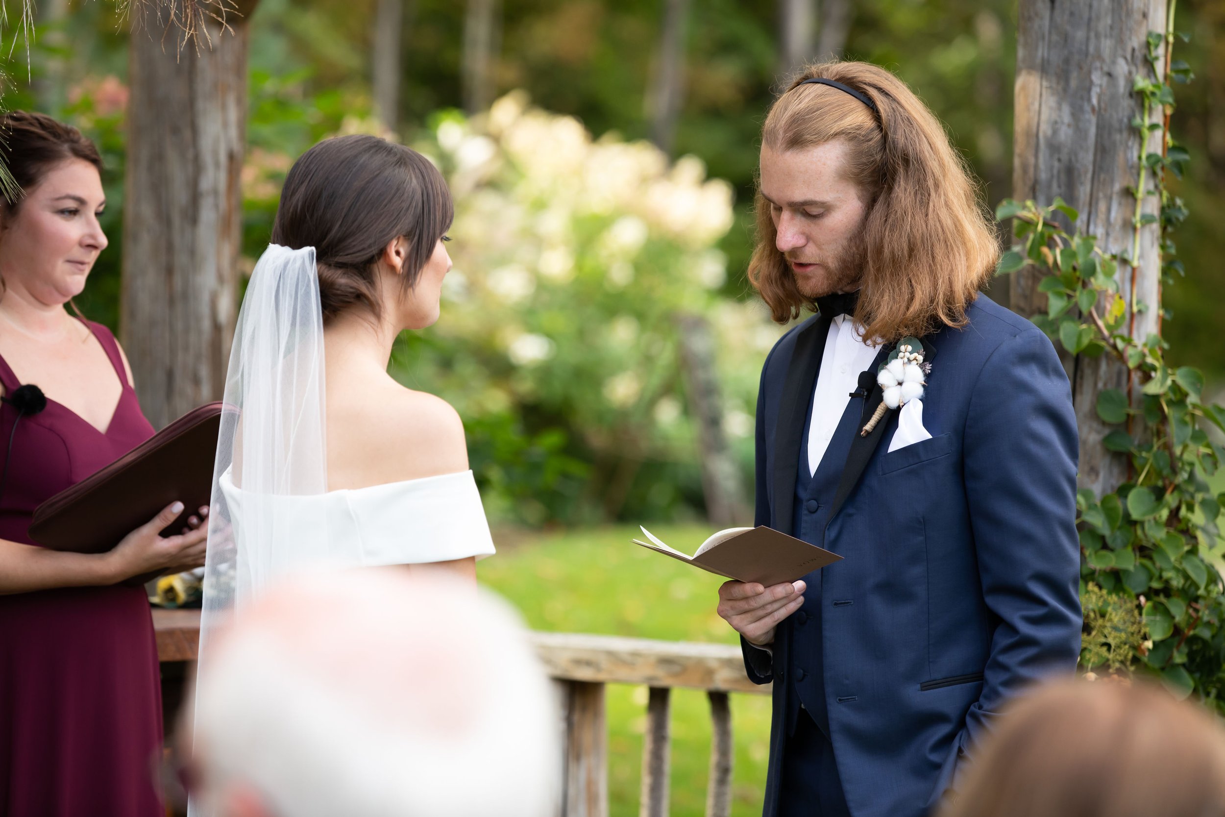 A couple getting married outdoors, with the officiant reading vows. The bride is in an off-shoulder dress and veil, the groom in a navy suit with boutonniere, and the officiant in a maroon dress holding a book.