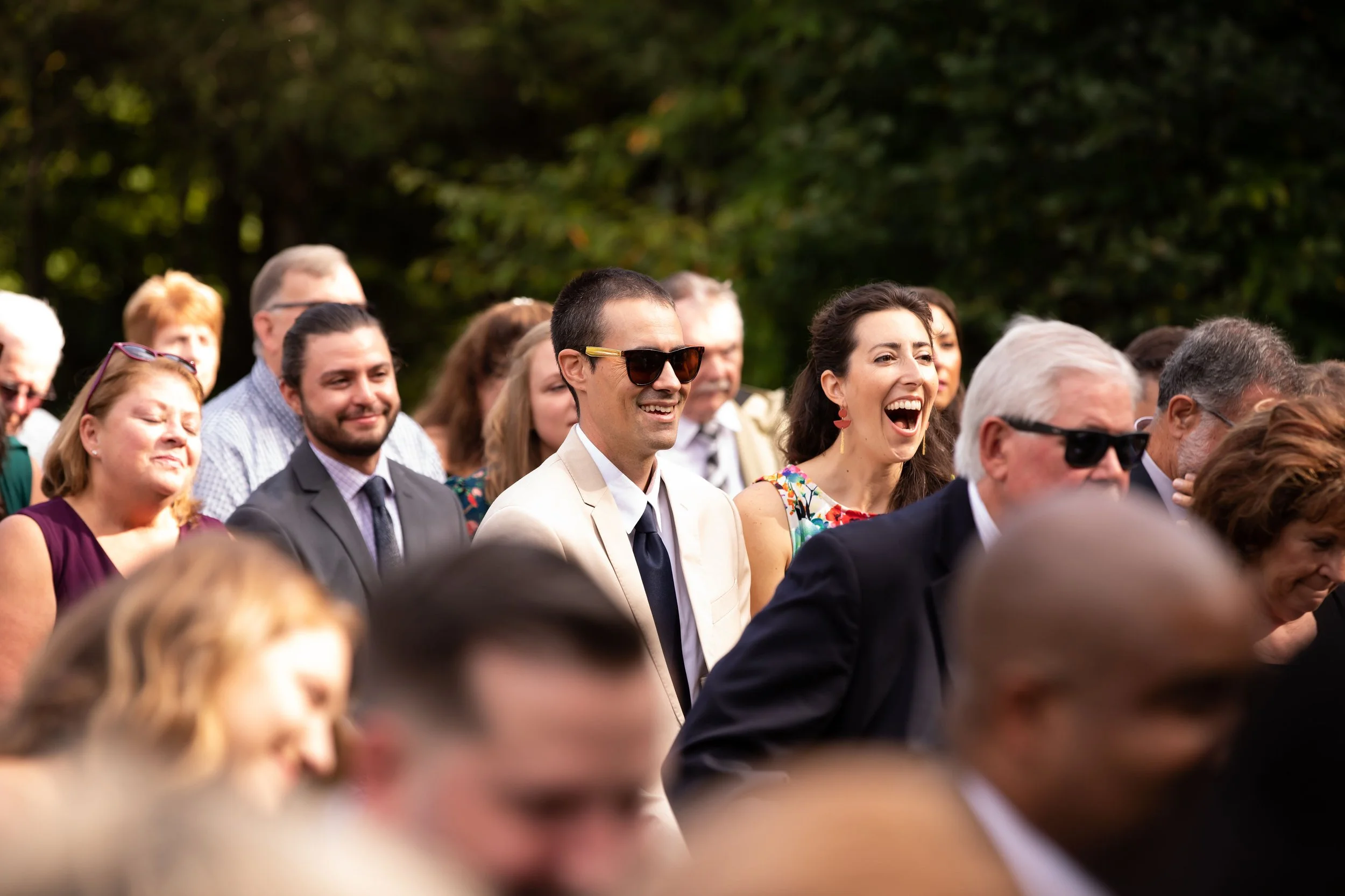 A group of people in formal attire, outdoors at a sunny event, smiling and laughing.