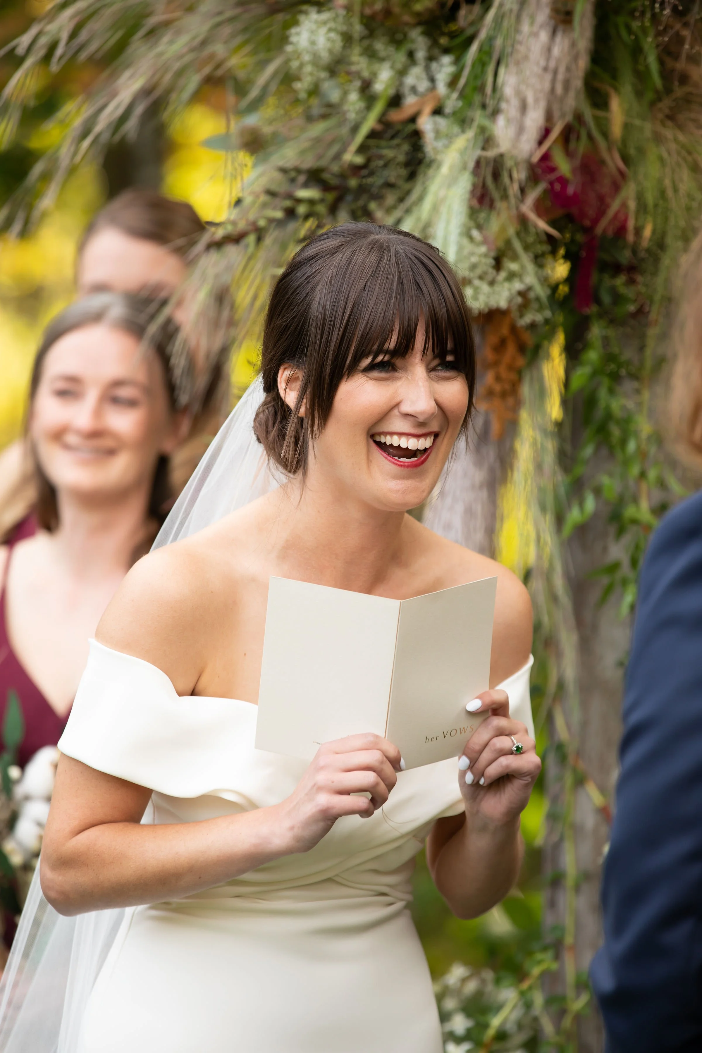 A bride with dark hair and a white off-shoulder wedding dress smiling and holding a vows booklet during her outdoor wedding ceremony, with a woman in a maroon dress and a man in a suit in the background, surrounded by greenery and floral decorations.