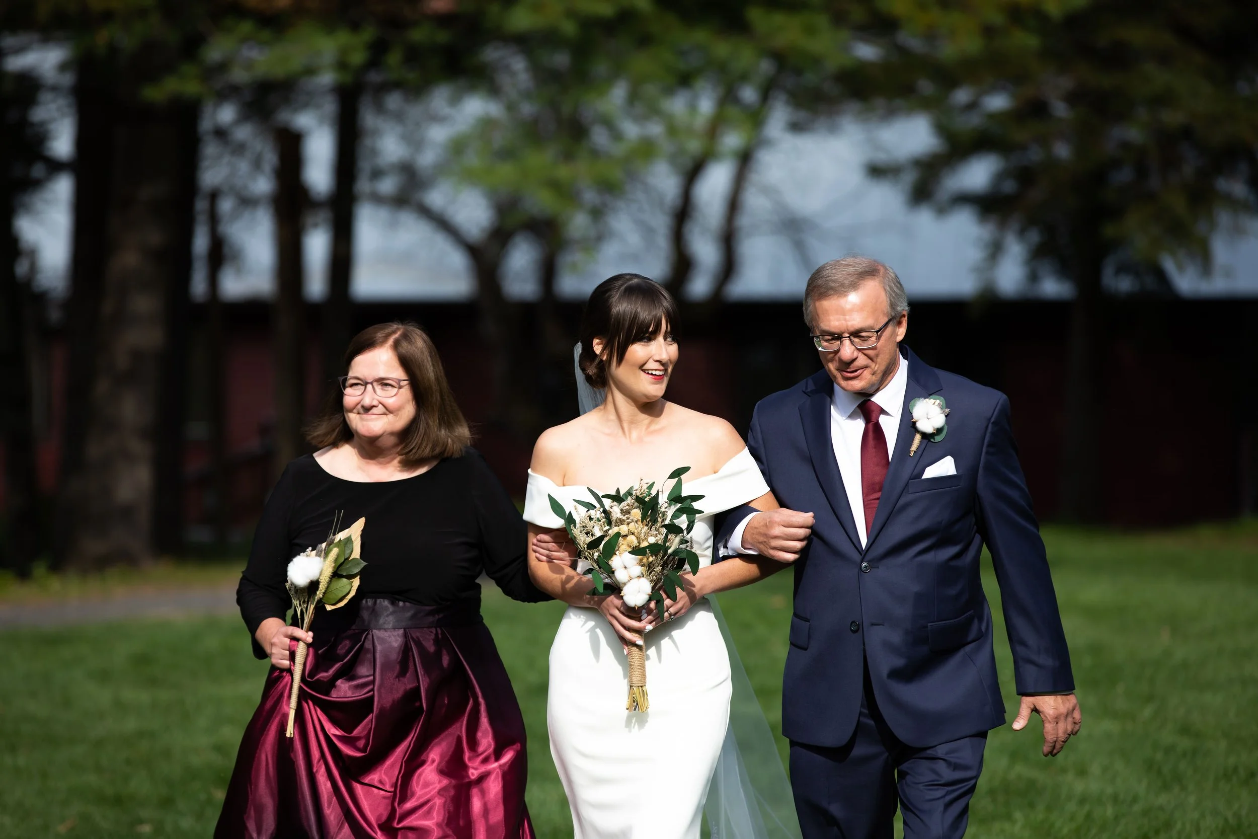 A bride in a white wedding dress holding a bouquet, walking arm-in-arm with her parents outdoors in a park or garden during her wedding.