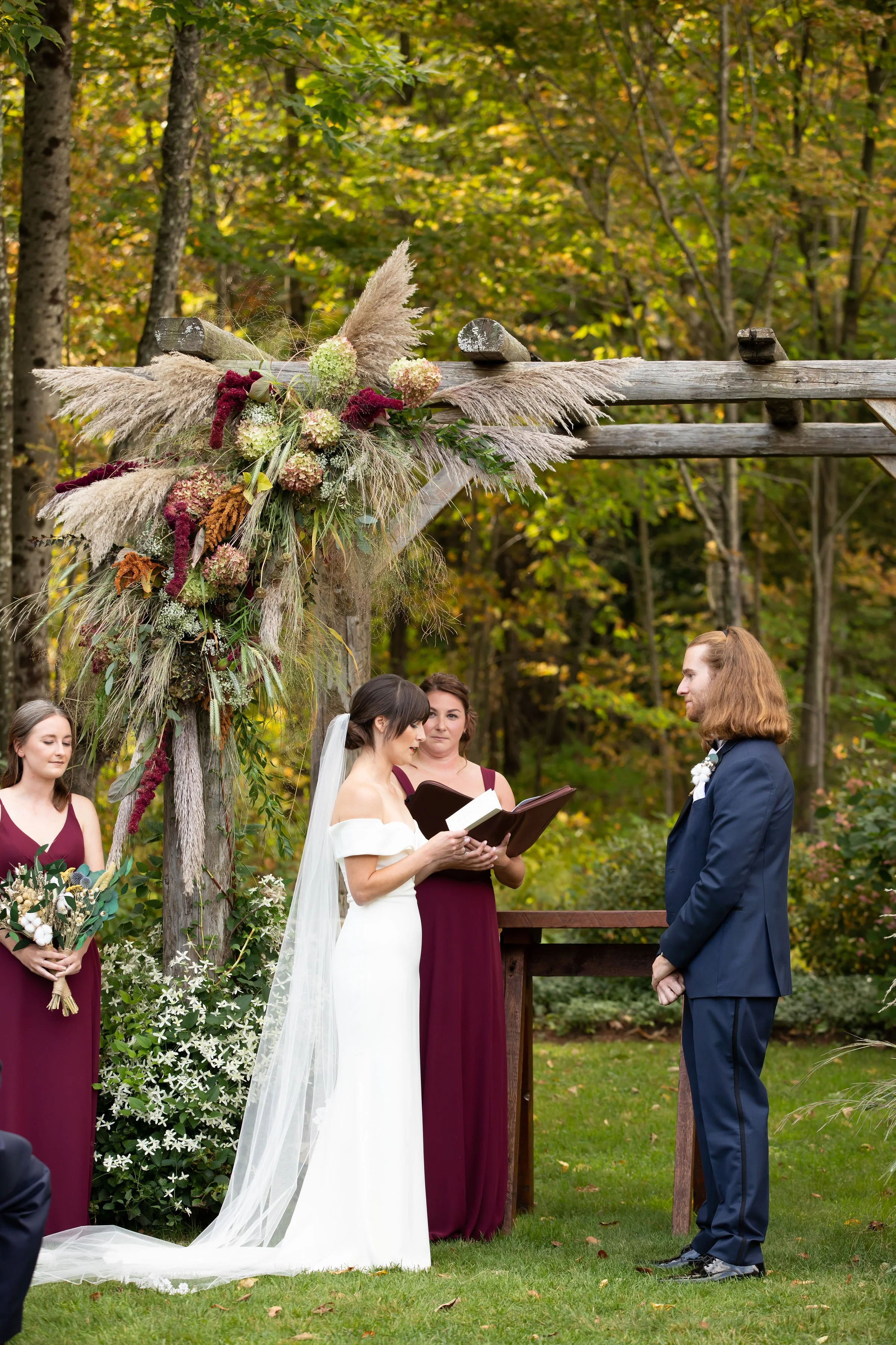 A wedding ceremony outdoors in a forest, with a bride and groom exchanging vows under a wooden arch decorated with pampas grass and flowers, officiated by a woman holding a book. Bridesmaids in maroon dresses stand nearby.