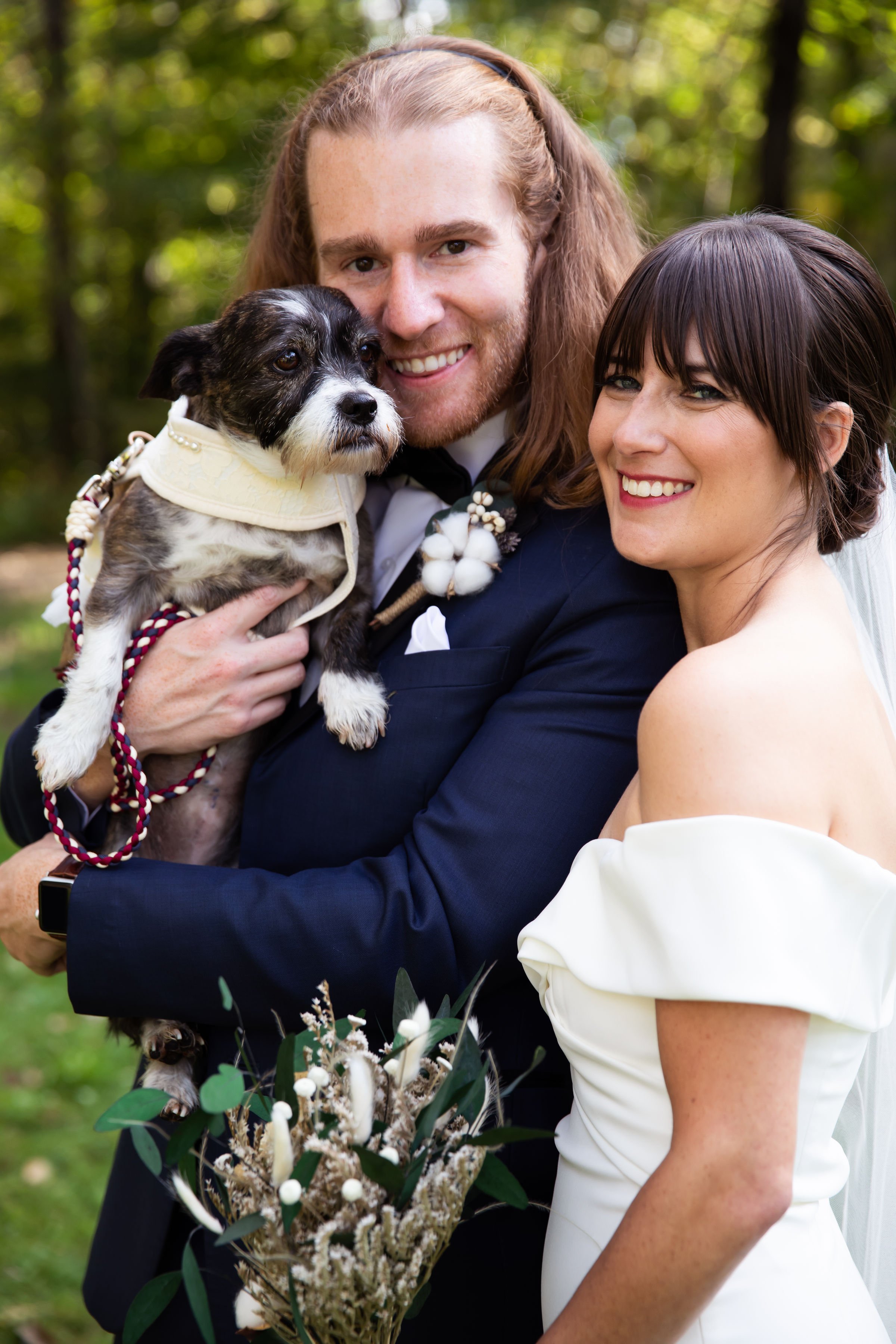 A newlywed couple holding a small dog outdoors, with green trees in the background. The groom is wearing a dark suit and the bride is in a white dress holding a bouquet of flowers.