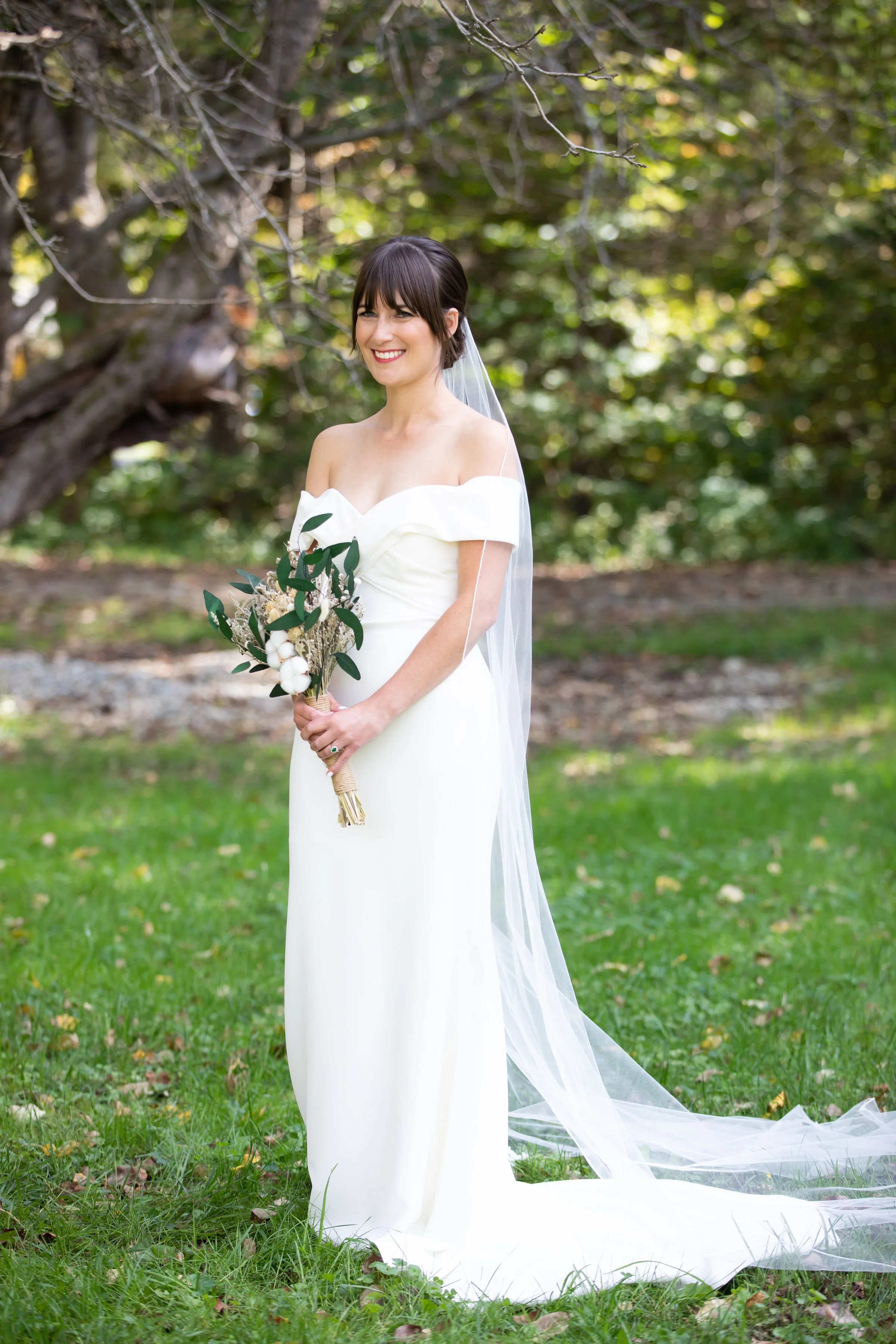 A smiling bride in a white off-shoulder wedding dress holding a bouquet of greenery and cotton flowers, standing outdoors on grass with a large tree in the background.