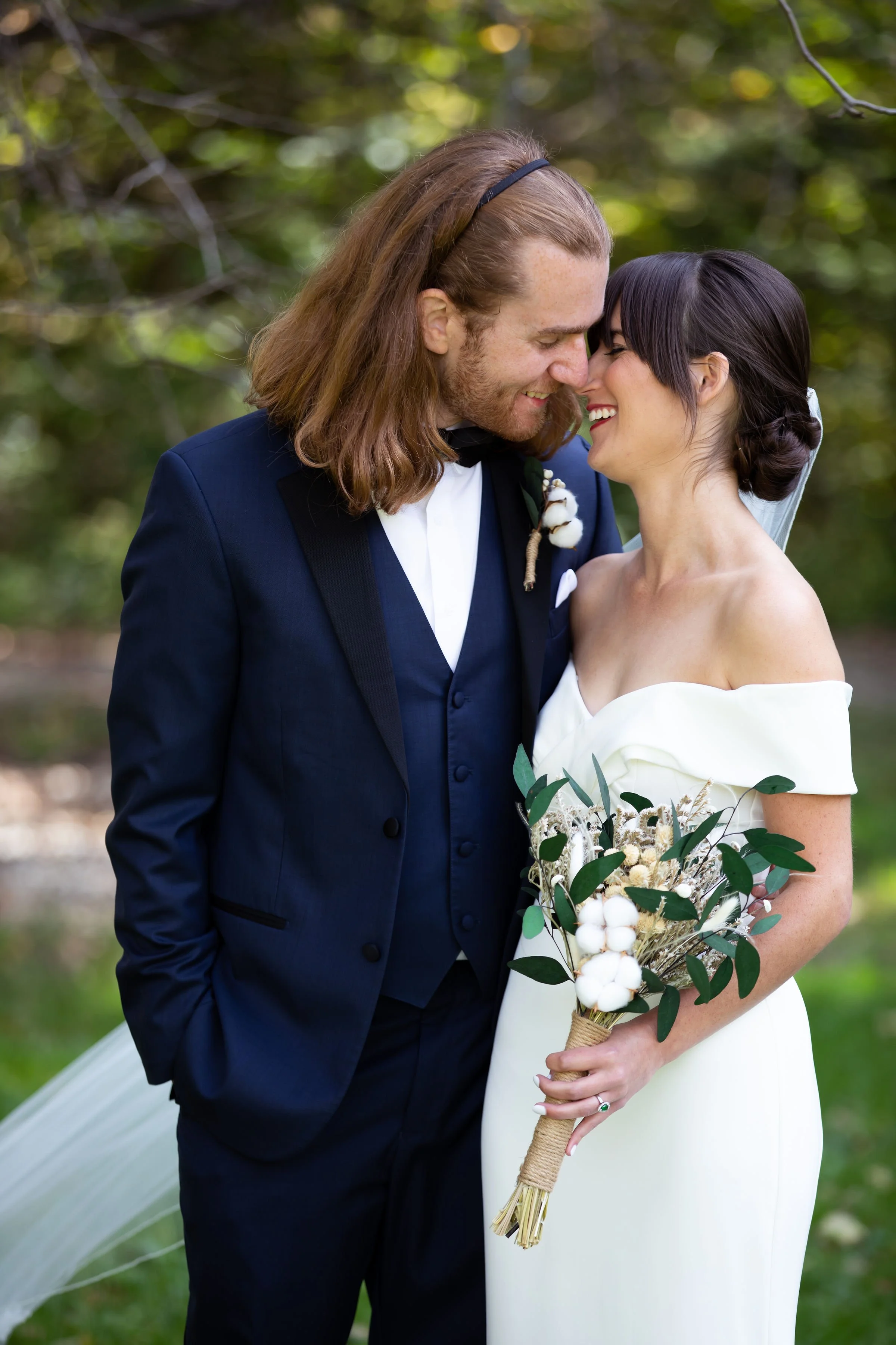 A newlywed couple, smiling and touching noses, stands outdoors in a garden or park setting. The groom has long, reddish-brown hair and is dressed in a dark blue tuxedo with a bow tie, holding his hands in his pockets. The bride has dark hair styled i