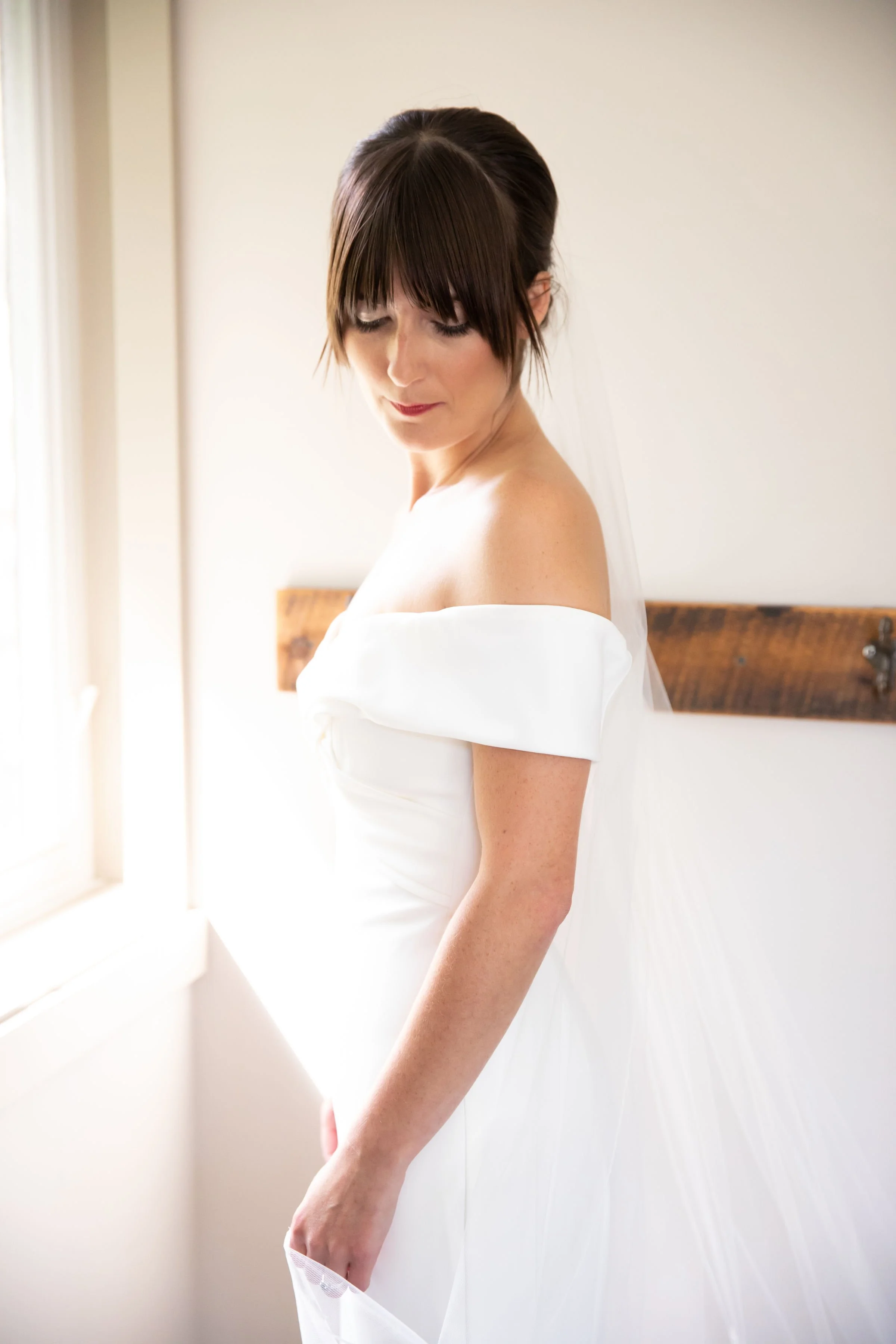 A bride wearing an off-shoulder white wedding dress, standing next to a window with natural light, with a wooden headboard visible behind her.