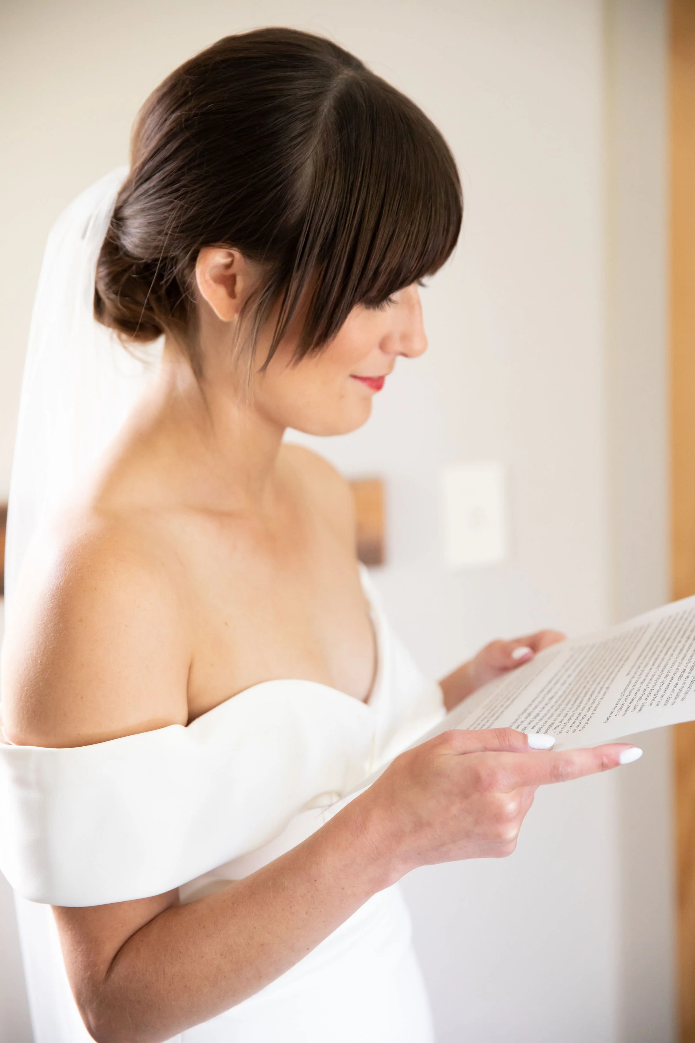A woman with brown hair styled in an updo, wearing a white off-the-shoulder dress, reading a piece of paper