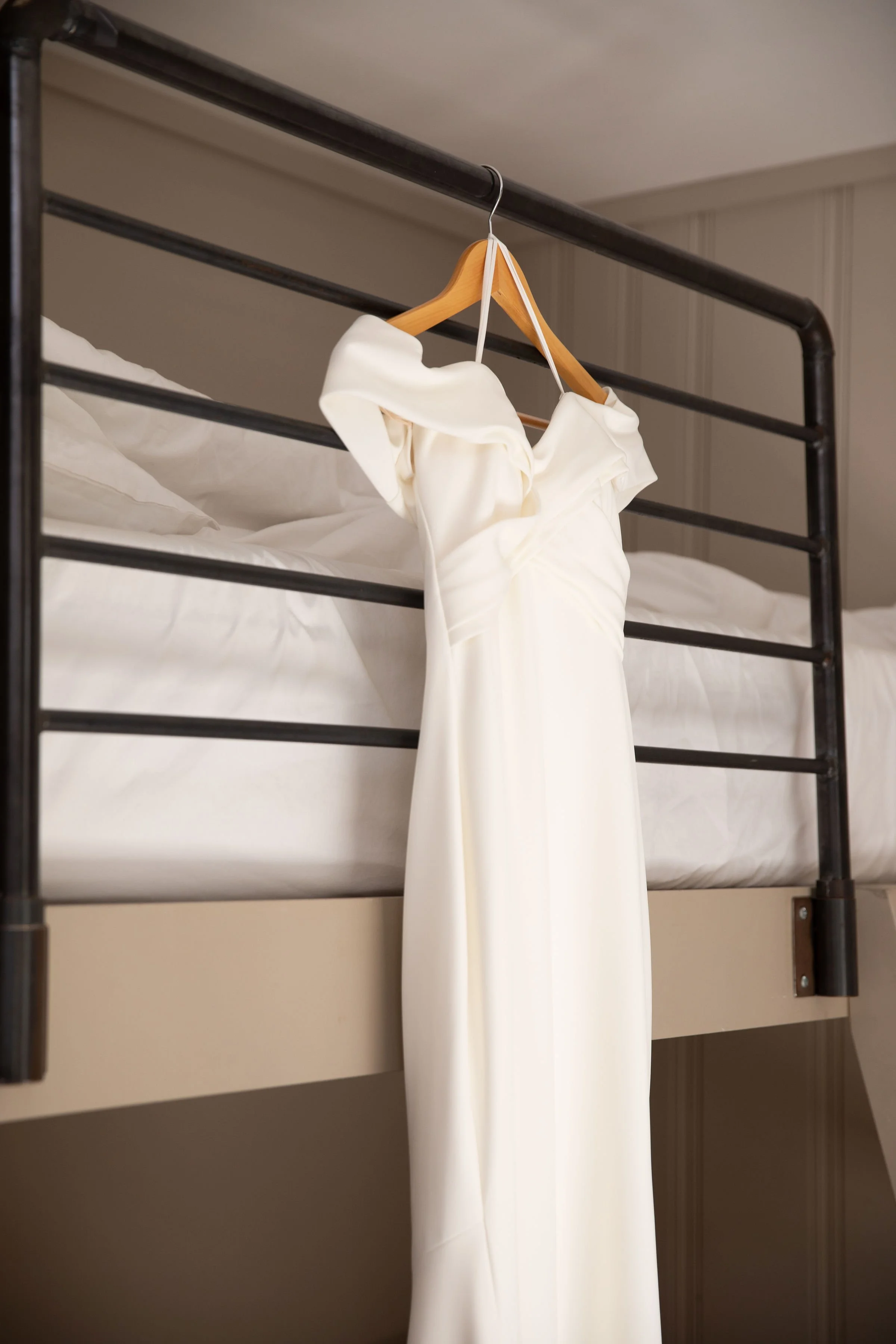 A white dress hangs on a wooden hanger from a metal bed frame.