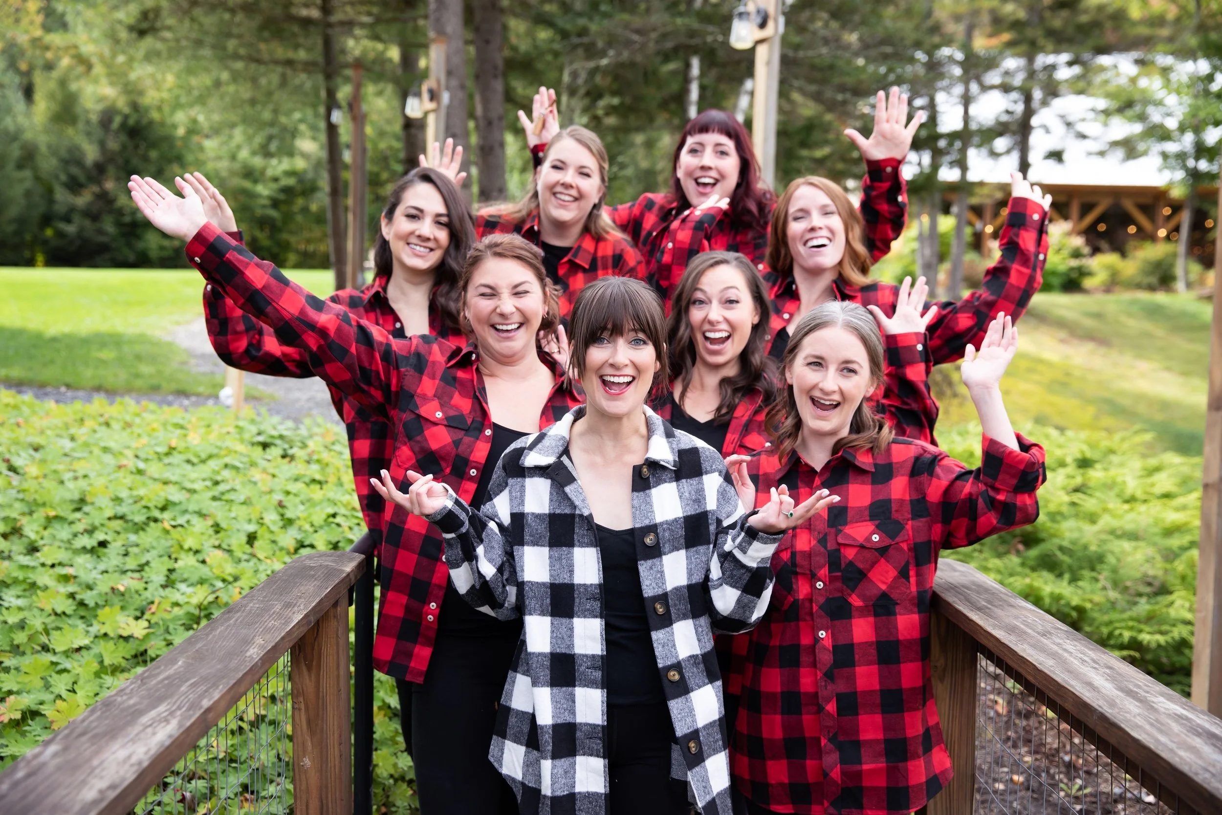 A group of women wearing red and black plaid shirts, smiling and posing outdoors on a wooden bridge surrounded by green trees.
