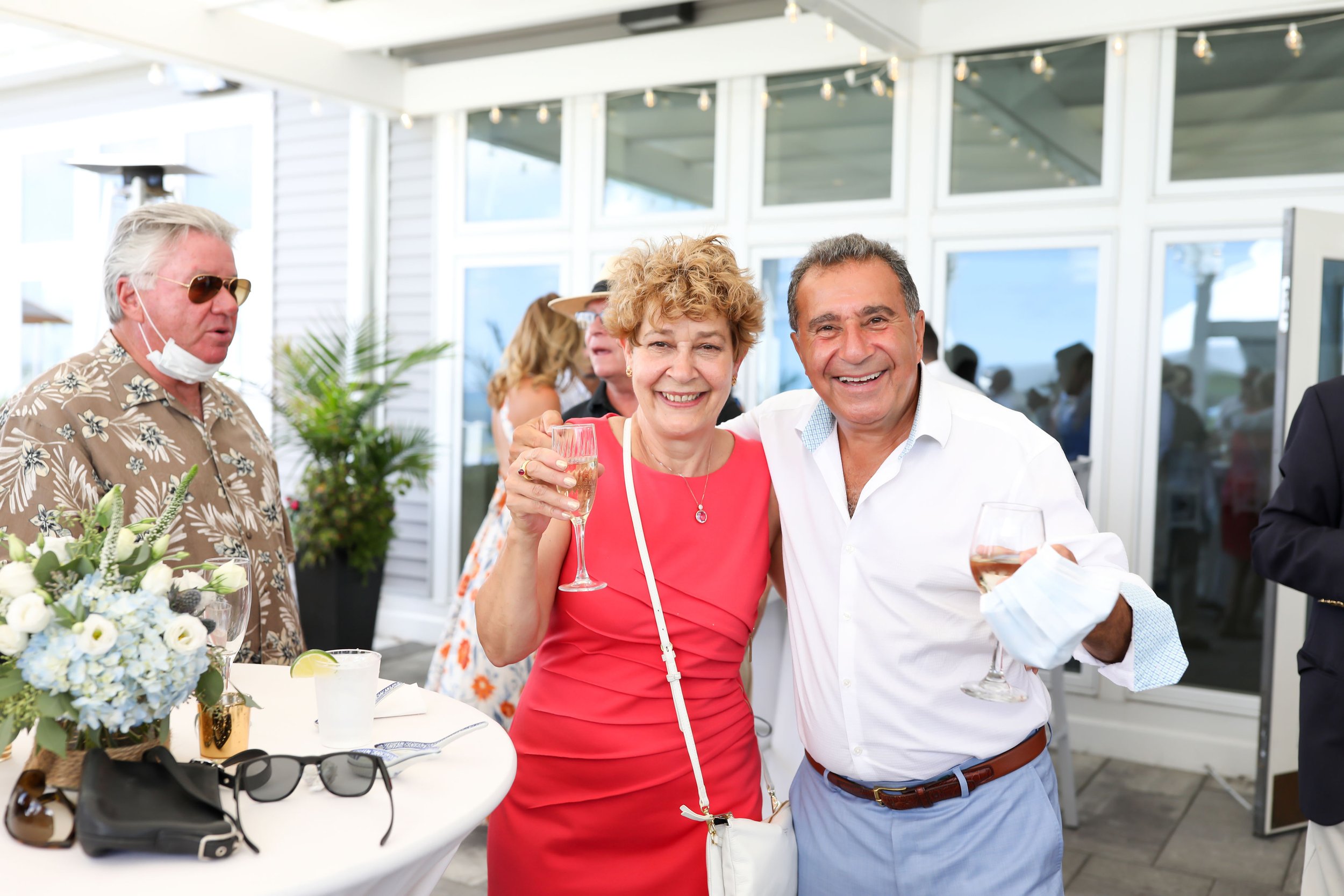 Two older adults, a woman in a pink dress and a man in a white shirt, smiling and holding glasses of wine at a social gathering, with other guests and decorations in the background.