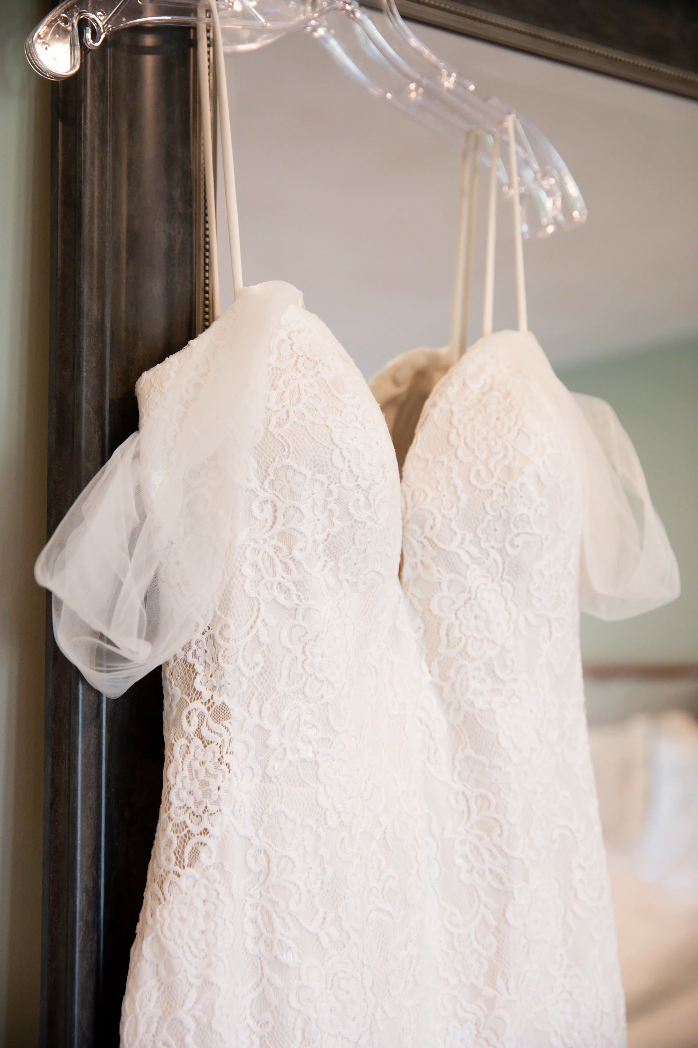 A white lace wedding dress hanging on a clear plastic hanger, displayed on a metal rack.