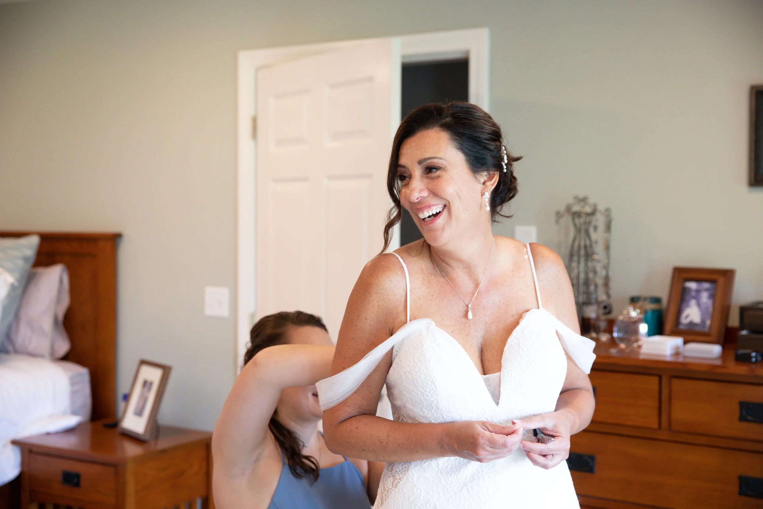 A joyful woman in a white dress with spaghetti straps and pearl jewelry, smiling as someone helps her with her dress in a bedroom.
