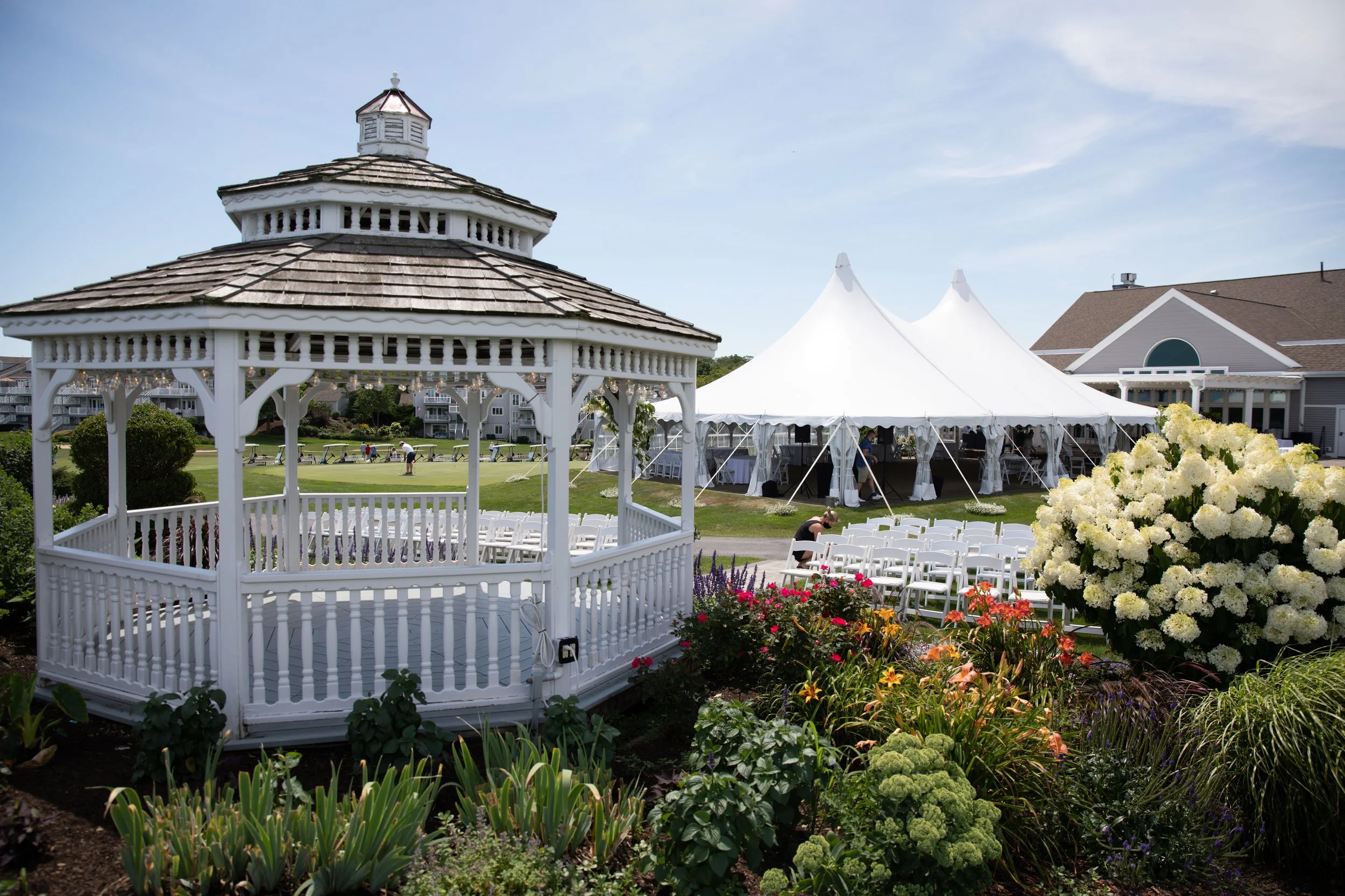A garden with a white gazebo, a white event tent, and rows of white chairs arranged for an outdoor event, surrounded by colorful flowers and greenery.