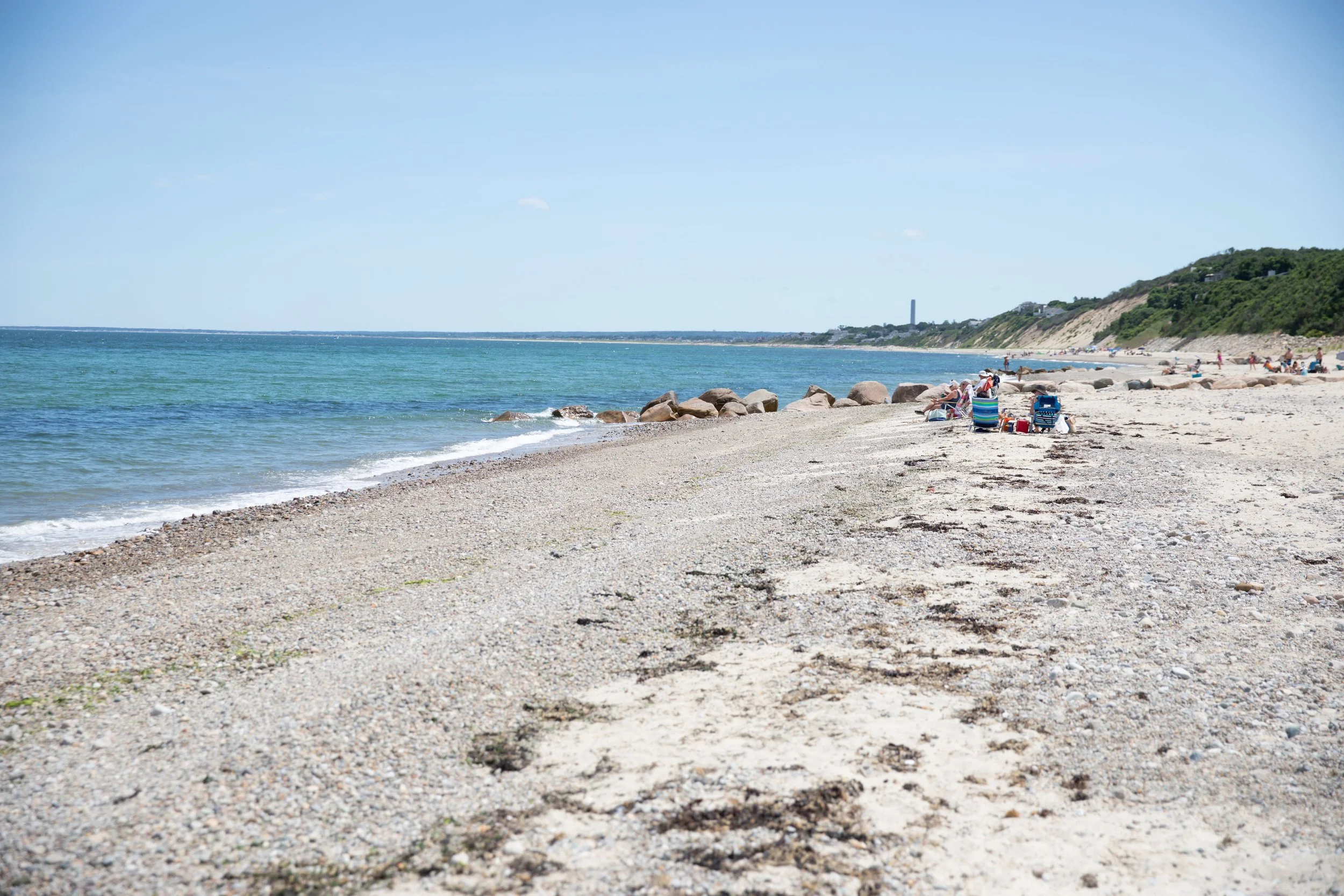 A sandy beach with people sitting on beach chairs and walking, near a calm blue ocean with a clear sky and some clouds.