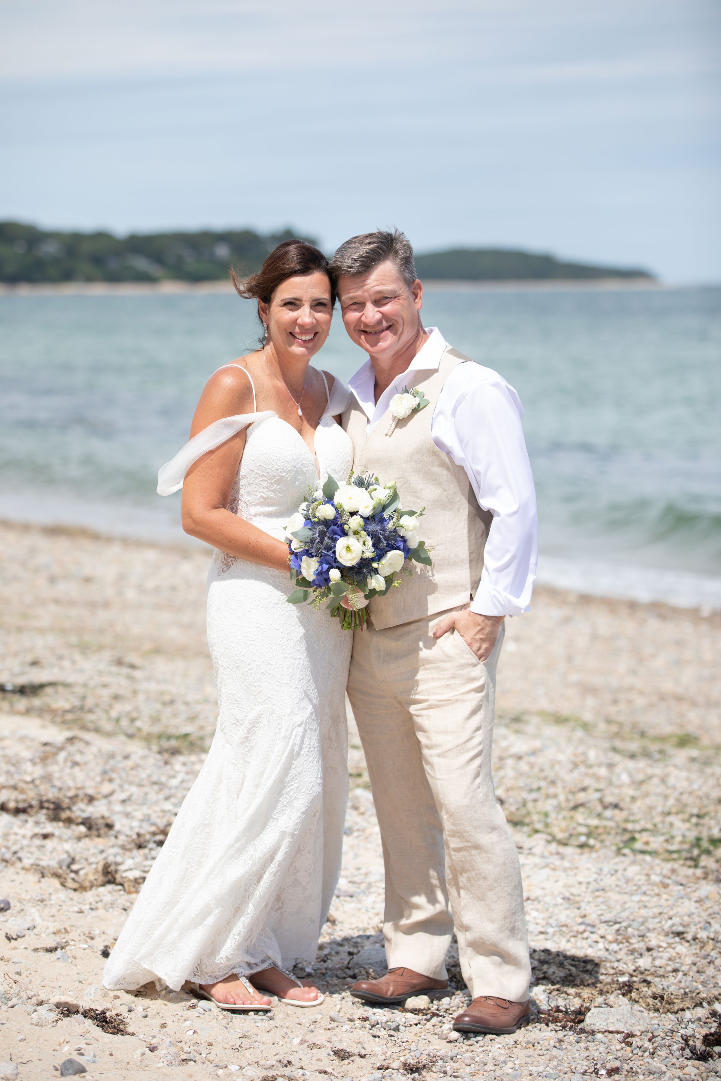 A newlywed couple stands on a beach, smiling, with the bride holding a bouquet of white and purple flowers. The bride wears a white lace wedding dress with off-the-shoulder sleeves, and the groom wears a white shirt with a beige vest and matching bei
