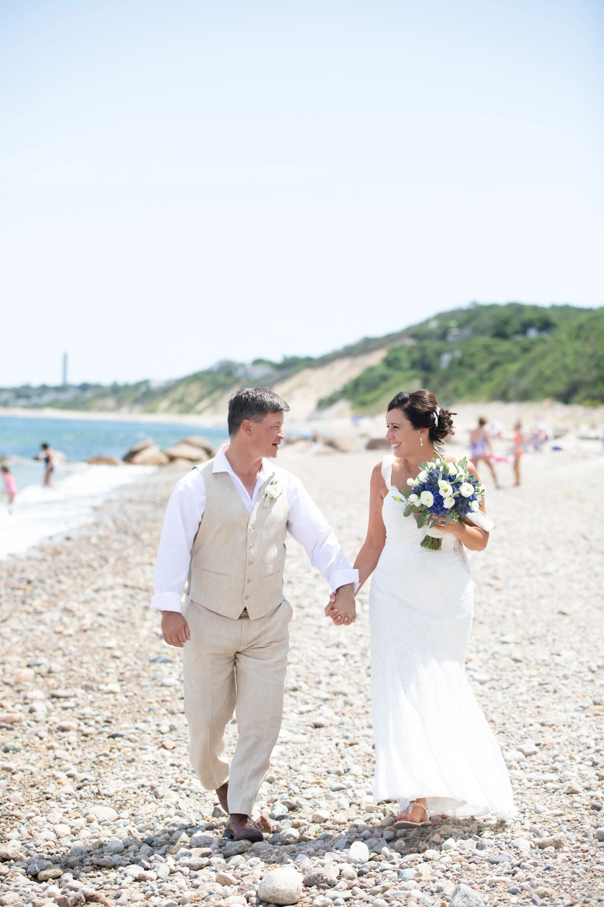 A bride and groom walking hand in hand on a pebble beach during their wedding, with the bride holding a bouquet of white and purple flowers and smiling at each other.