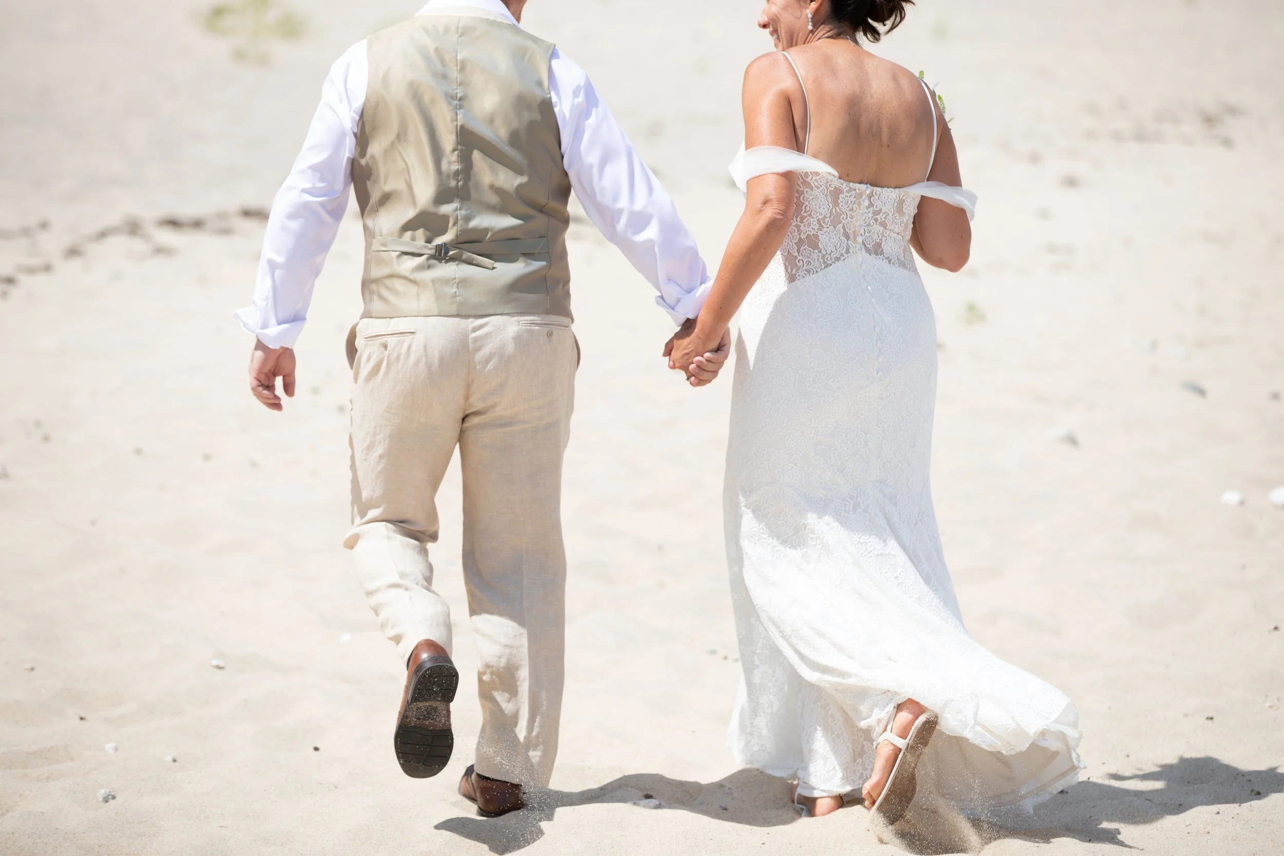 A couple dressed in wedding attire walking hand in hand on a sandy beach.