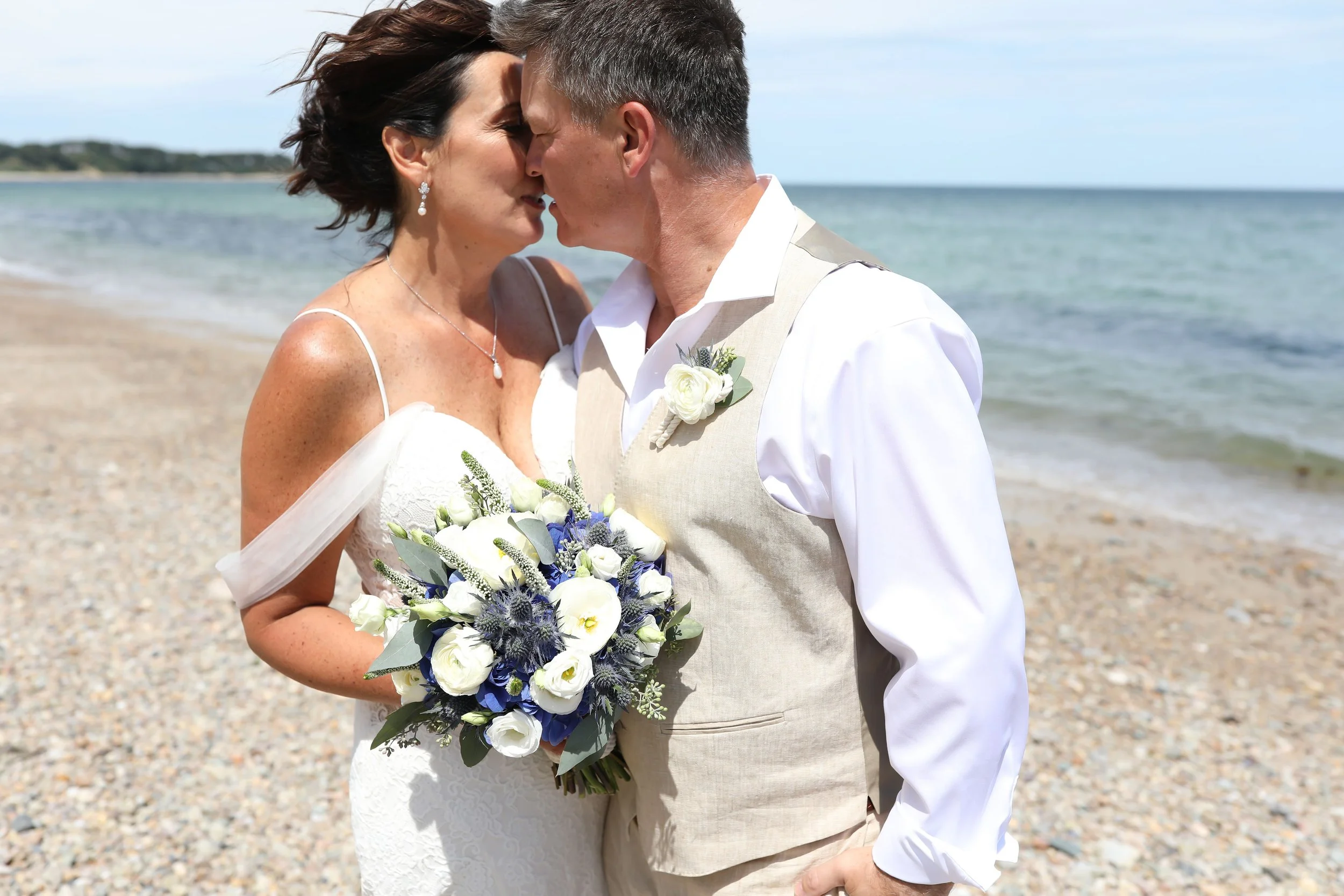 A bride and groom kiss on a beach, with the ocean and sky in the background. The bride holds a white and blue bouquet, and both are dressed in wedding attire.