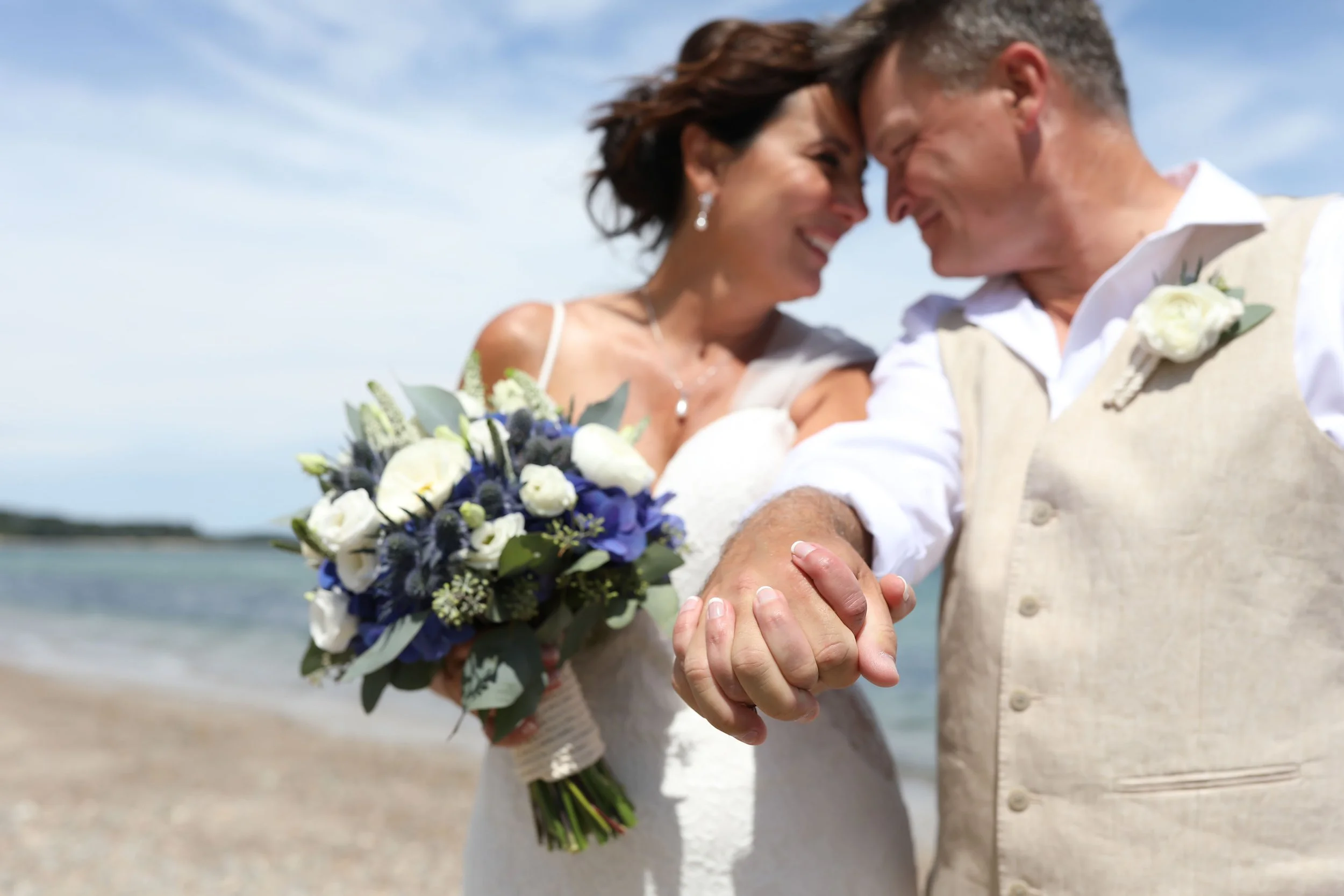 A happy couple in wedding attire on the beach, holding hands with their foreheads touching and smiling at each other, with the bride holding a bouquet of white and purple flowers.