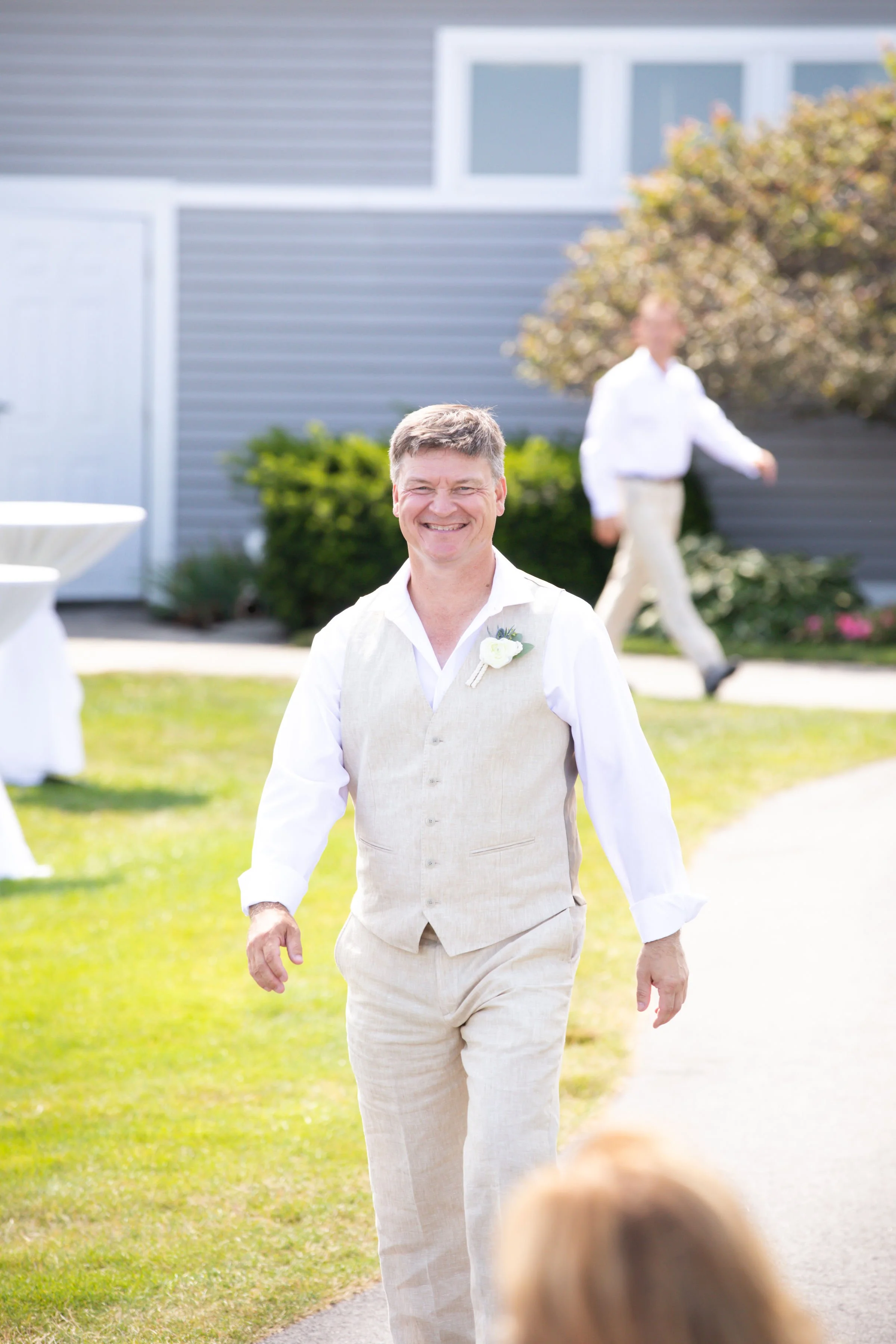 A smiling man in a beige vest and pants walking outdoors on a sunny day at a gathering, with another person blurred in the background and a house with gray siding behind him.